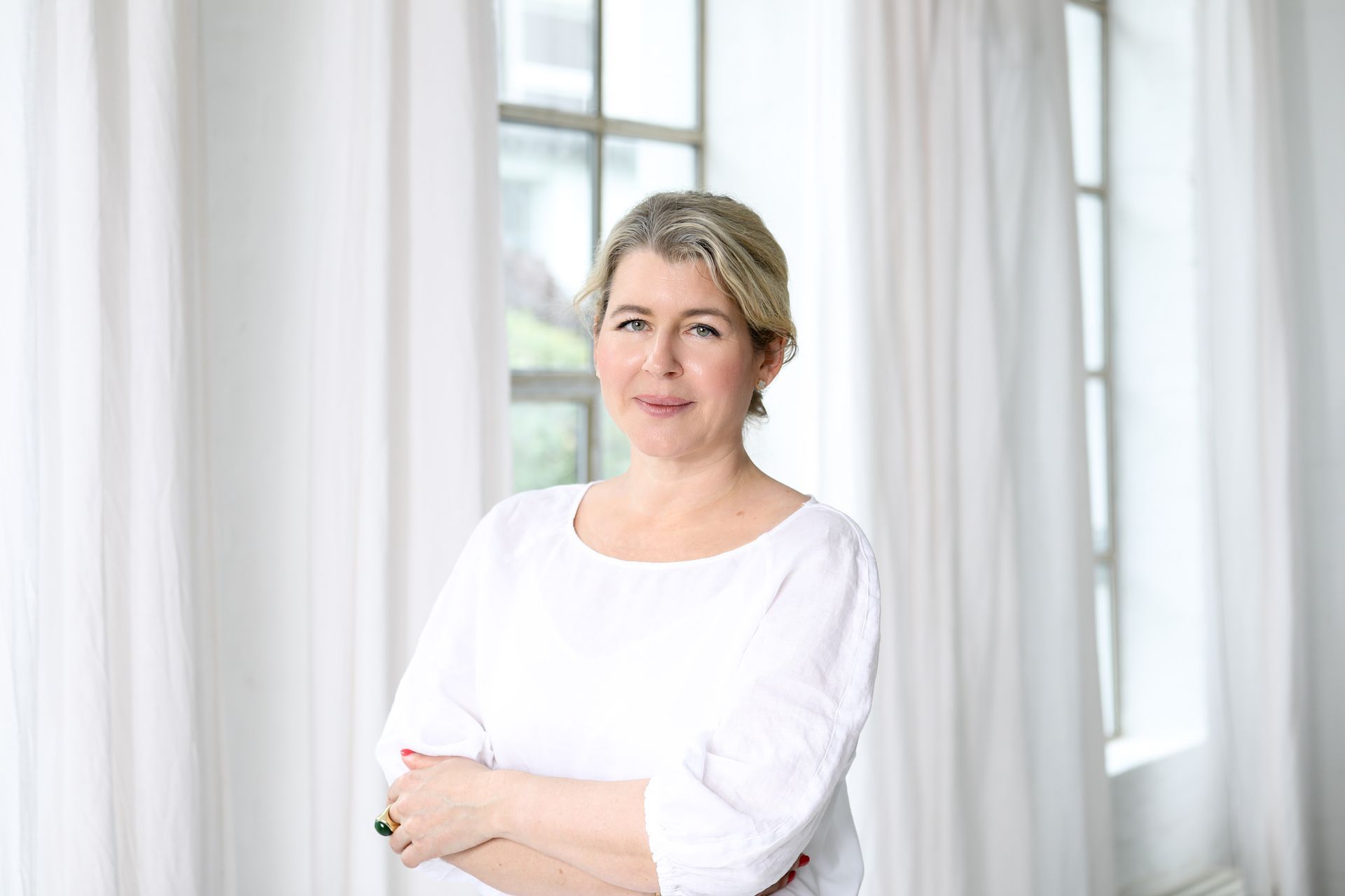 Eine Frau in einem weißen Hemd steht mit verschränkten Armen vor einem Fenster.