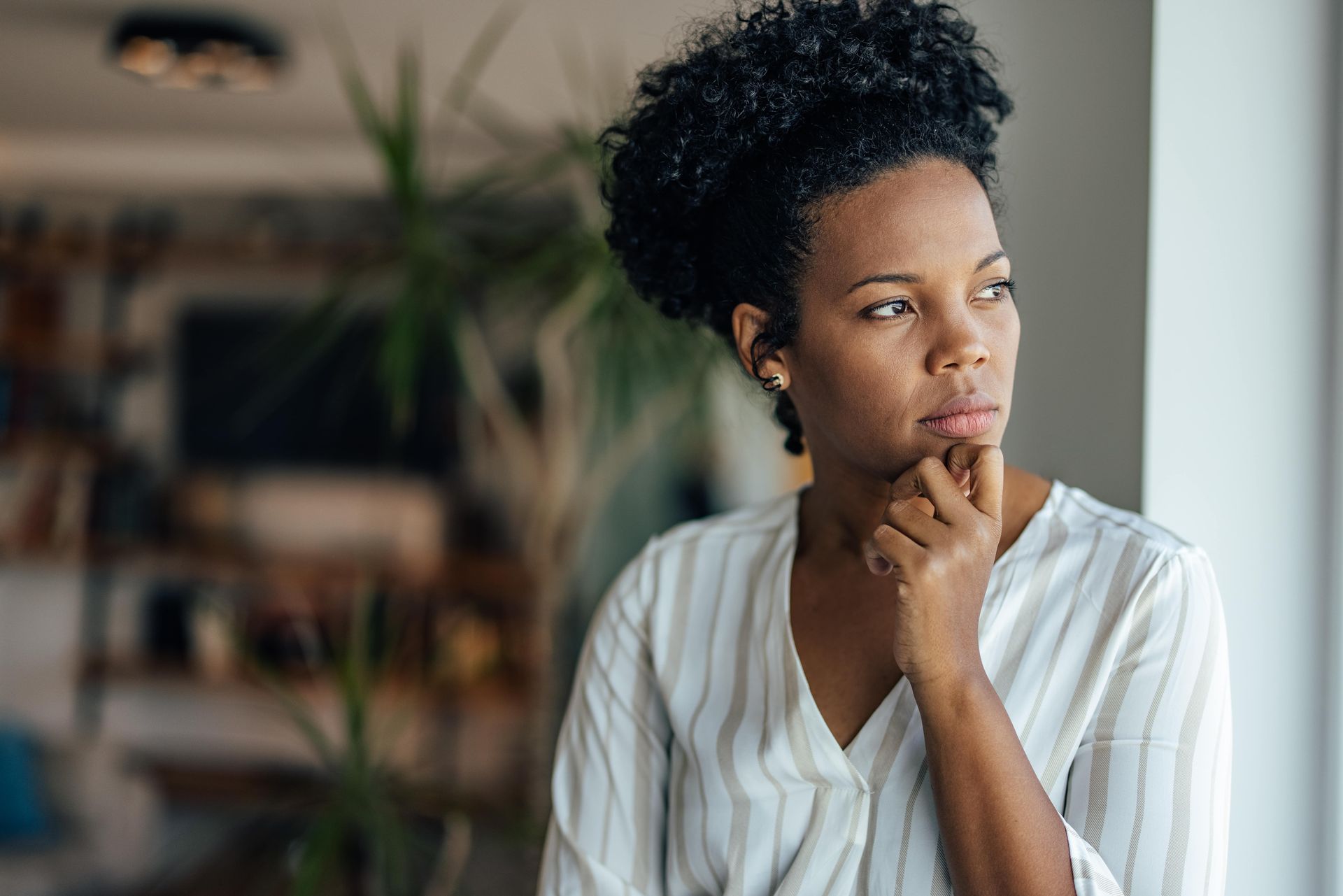 Woman looking out a window, hand on chin, thoughtful expression, bright room with a plant.