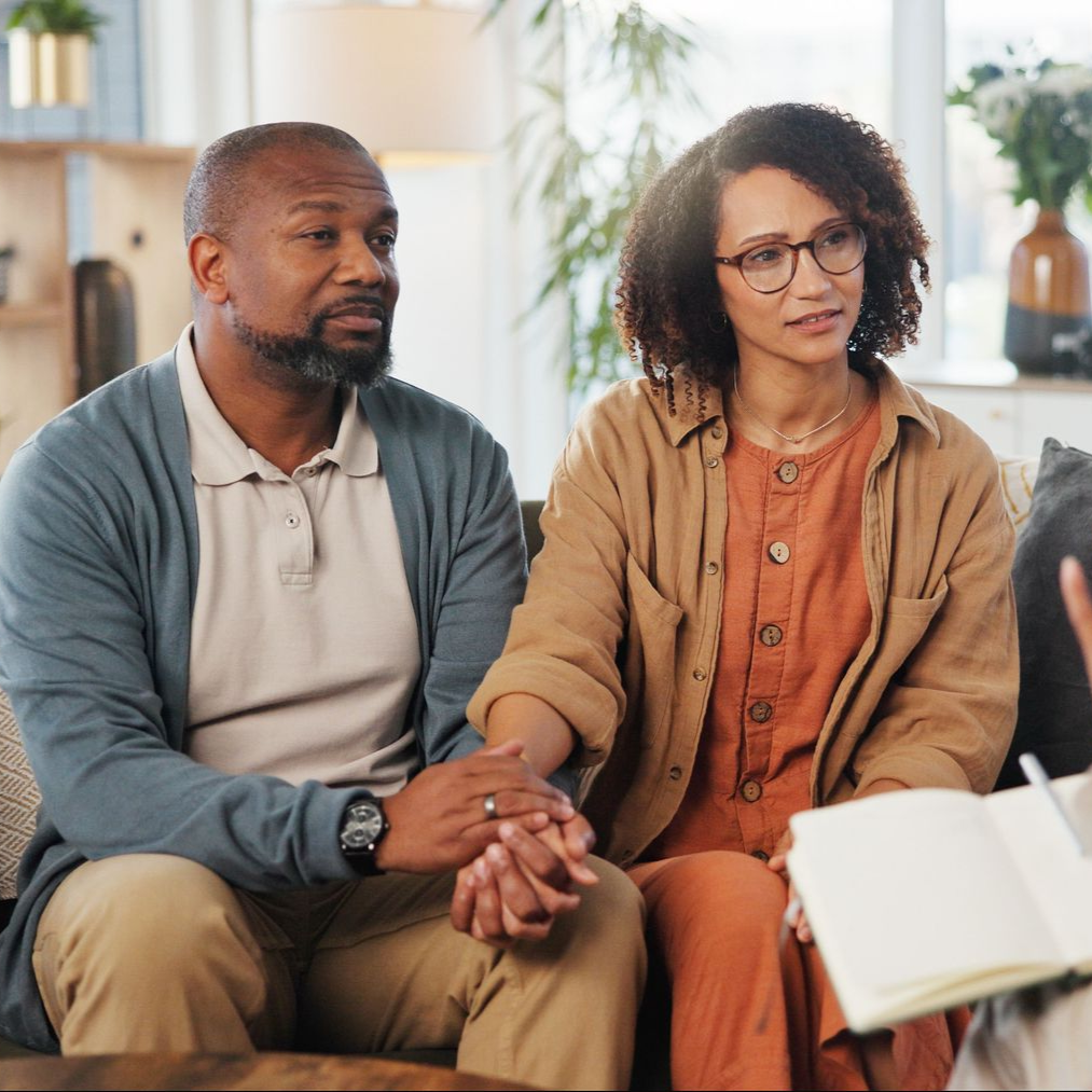 A therapist talking to a couple on a couch, in a well-lit living room setting.
