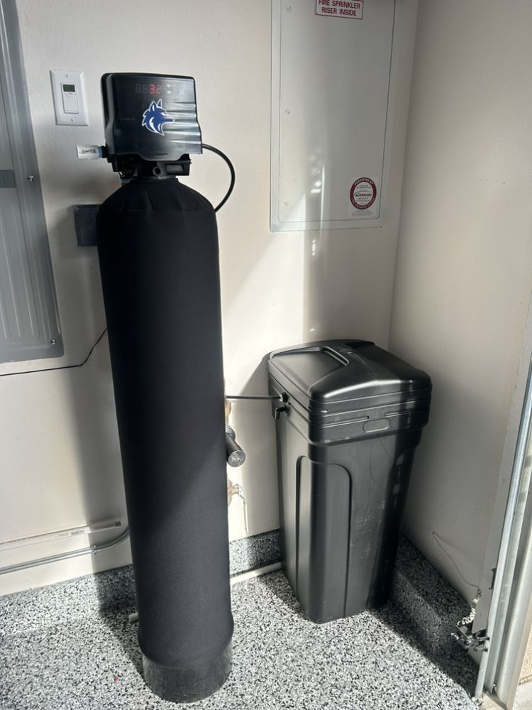 Water filtration system with metal tanks, a blue filter, and a white storage container against a gray wall.