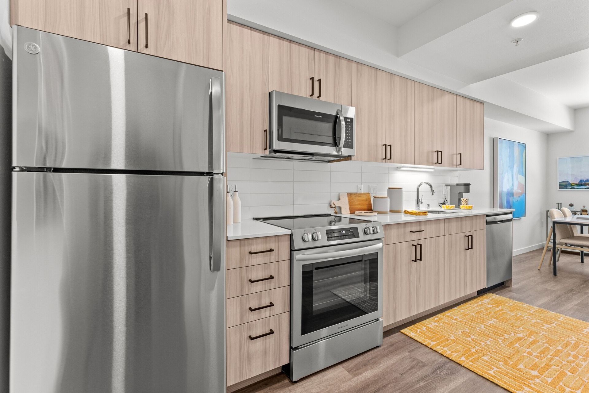 Modern kitchen with stainless steel appliances, light wood cabinets, and yellow rug.