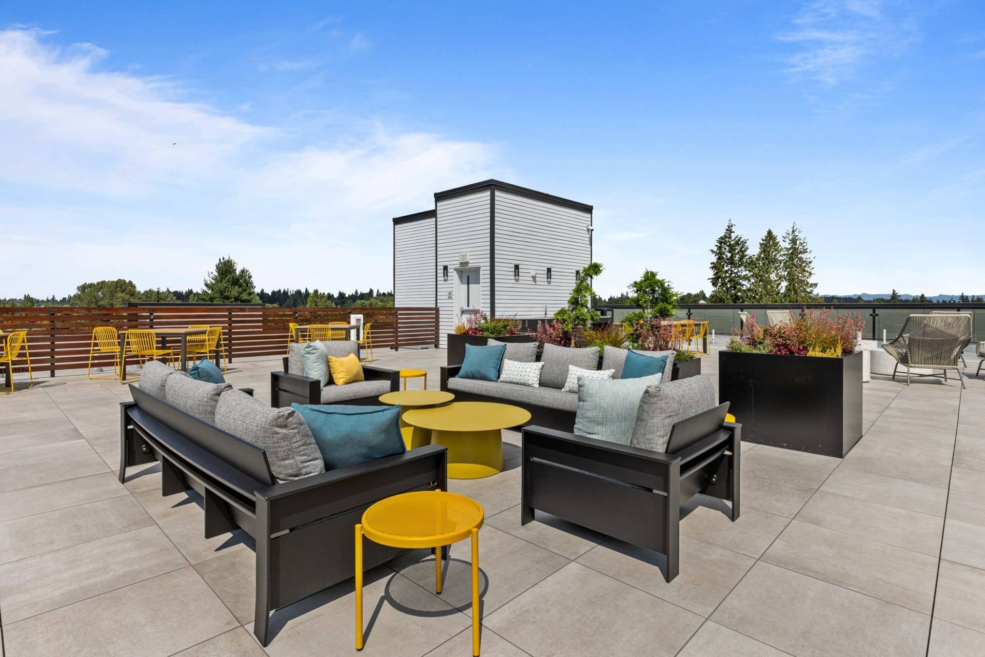 Rooftop patio with gray couches, yellow tables, and potted plants on a sunny day.