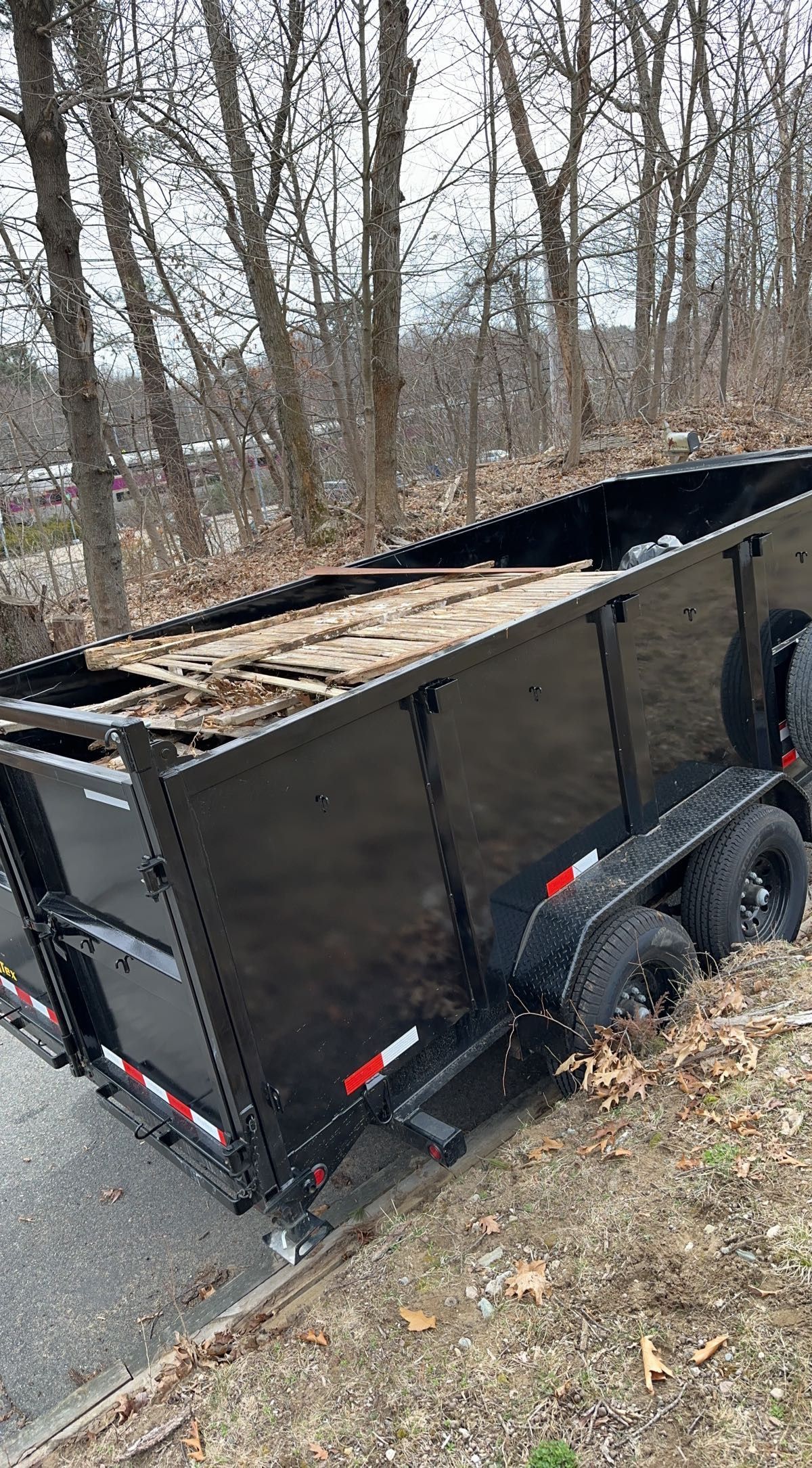 Black dump trailer filled with wood debris on a roadside with trees in the background.
