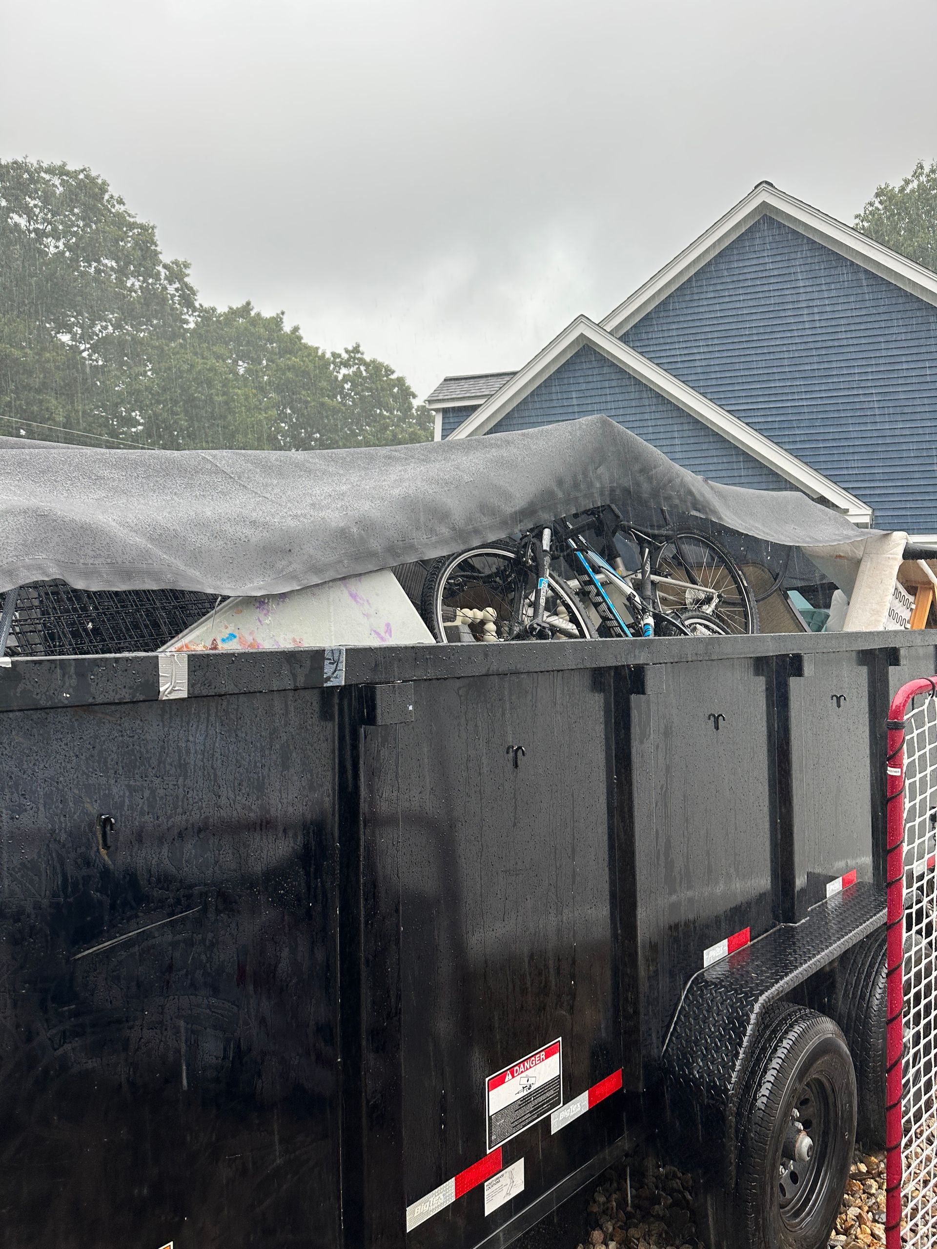 Black dumpster filled with debris, backed by a house with blue shingles, gray sky.
