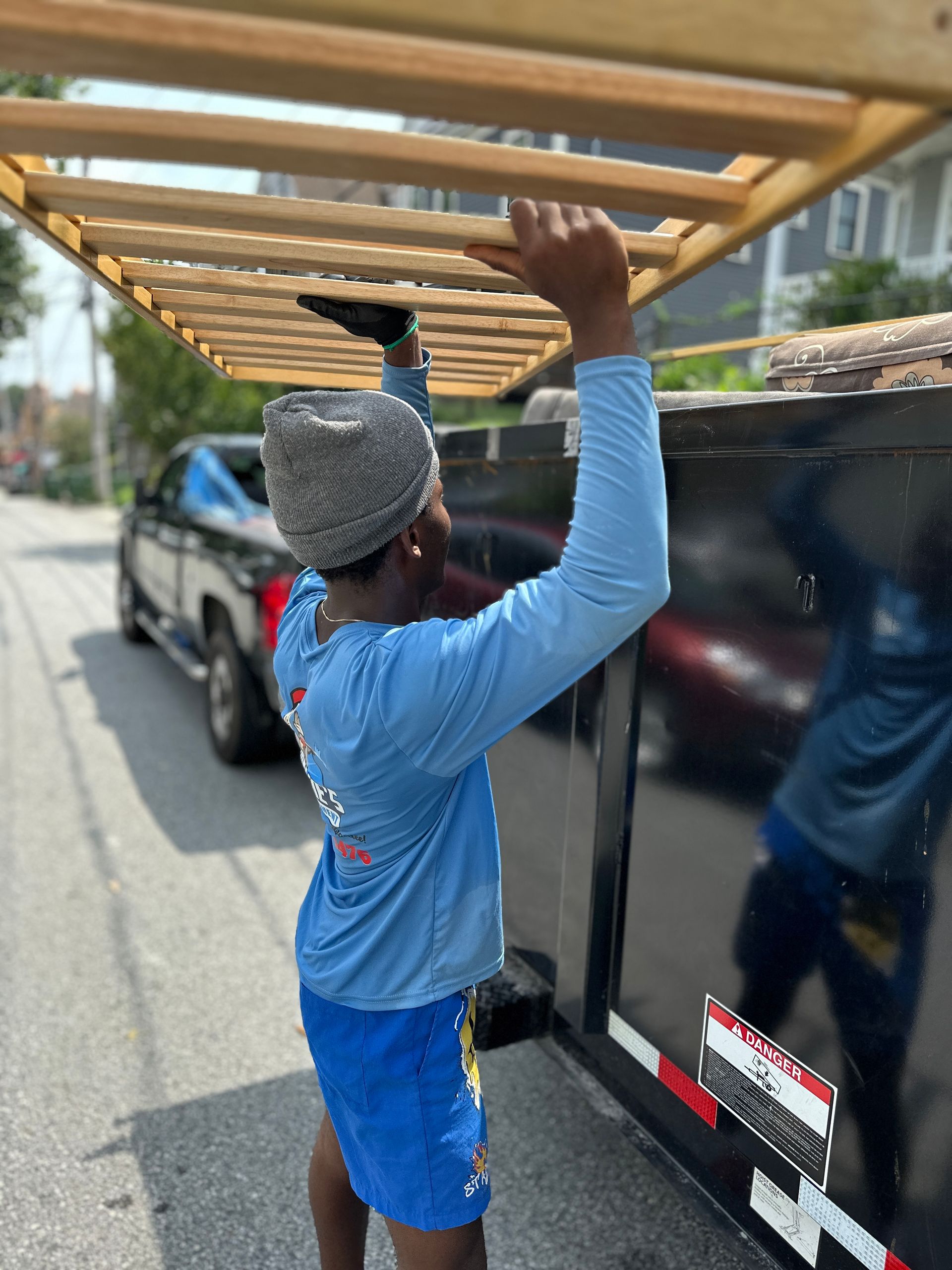 Man in blue shirt and shorts lifting wooden structure onto a black trailer on a sunny street.