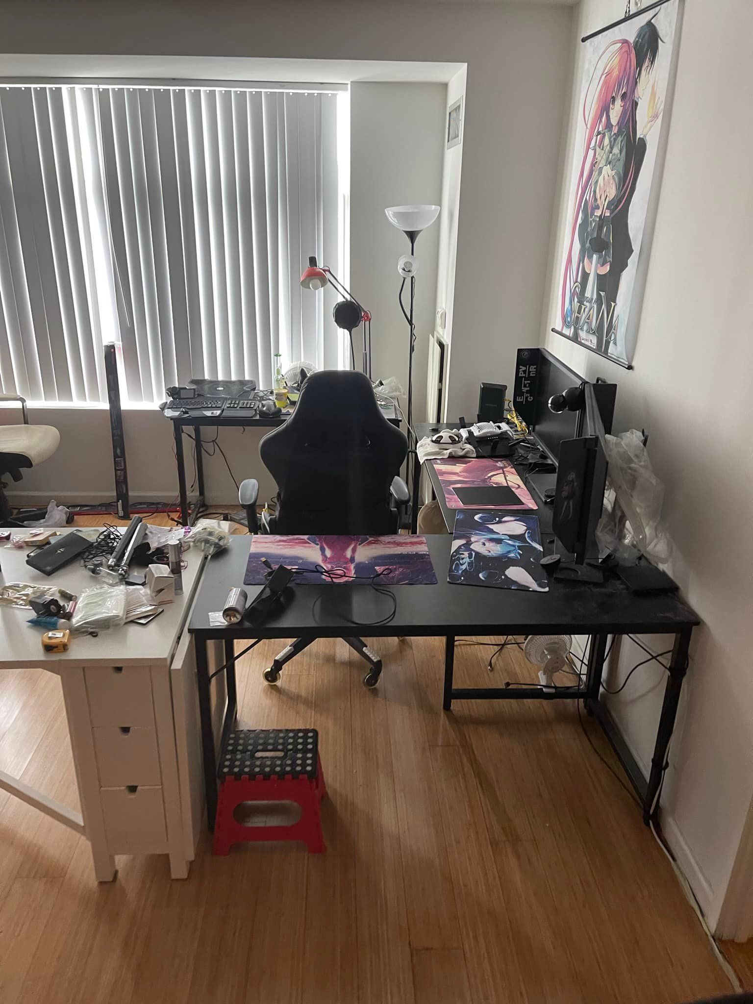 A cluttered home office with a gaming desk, computer, chair, and artwork. Light from a window illuminates the room.