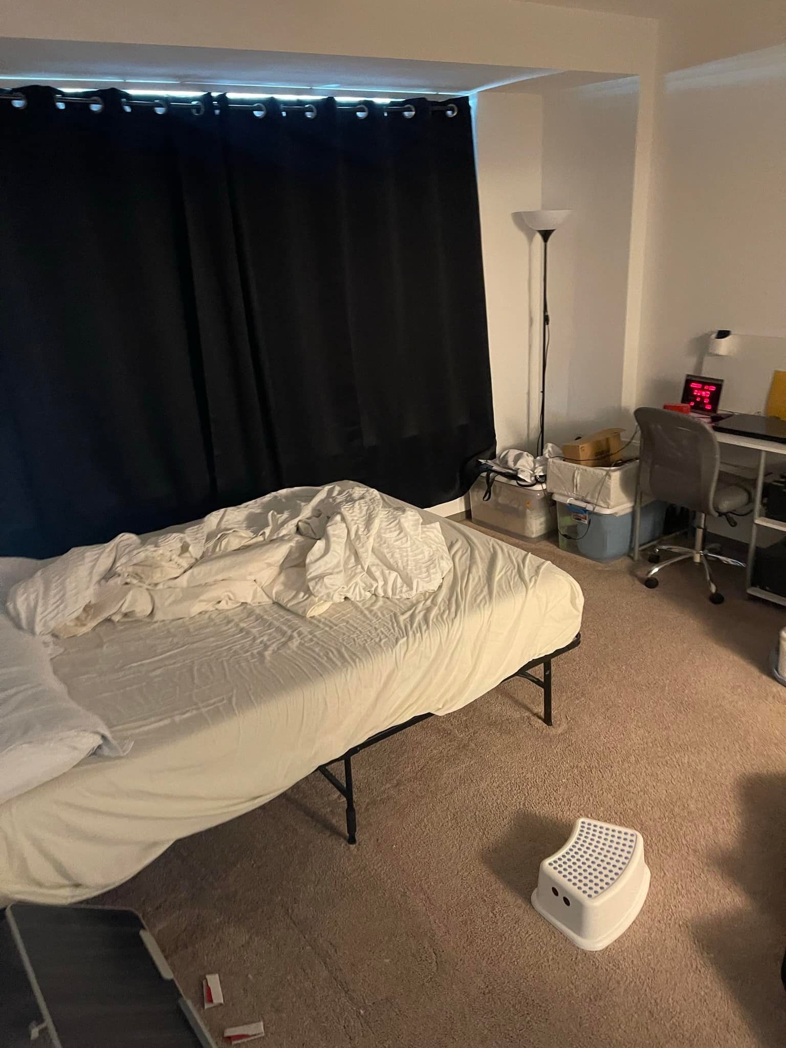 Bedroom with unmade bed, black curtains, desk, floor lamp, and small white step stool.