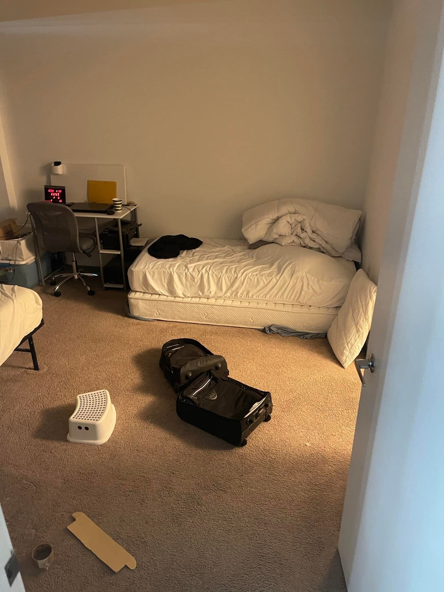 Bedroom with unmade bed, desk, open luggage, and various items on the carpeted floor.