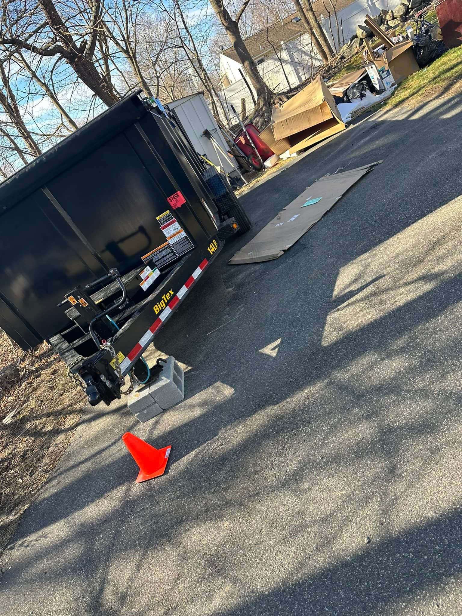 Black trailer angled in driveway, orange cone, cardboard and debris in background.