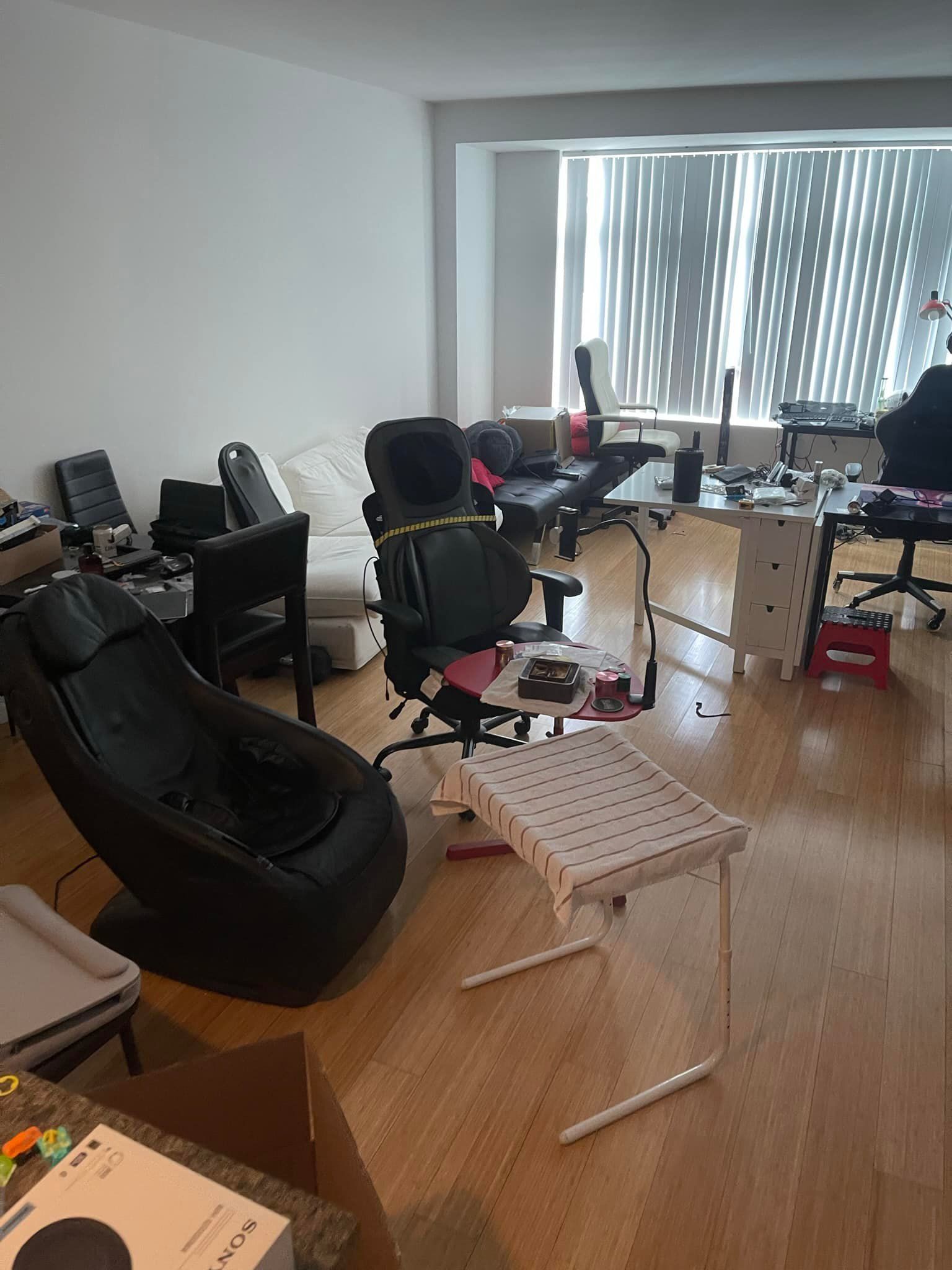 A cluttered living room with wooden floors. Various chairs, desks, and equipment are scattered around the room.