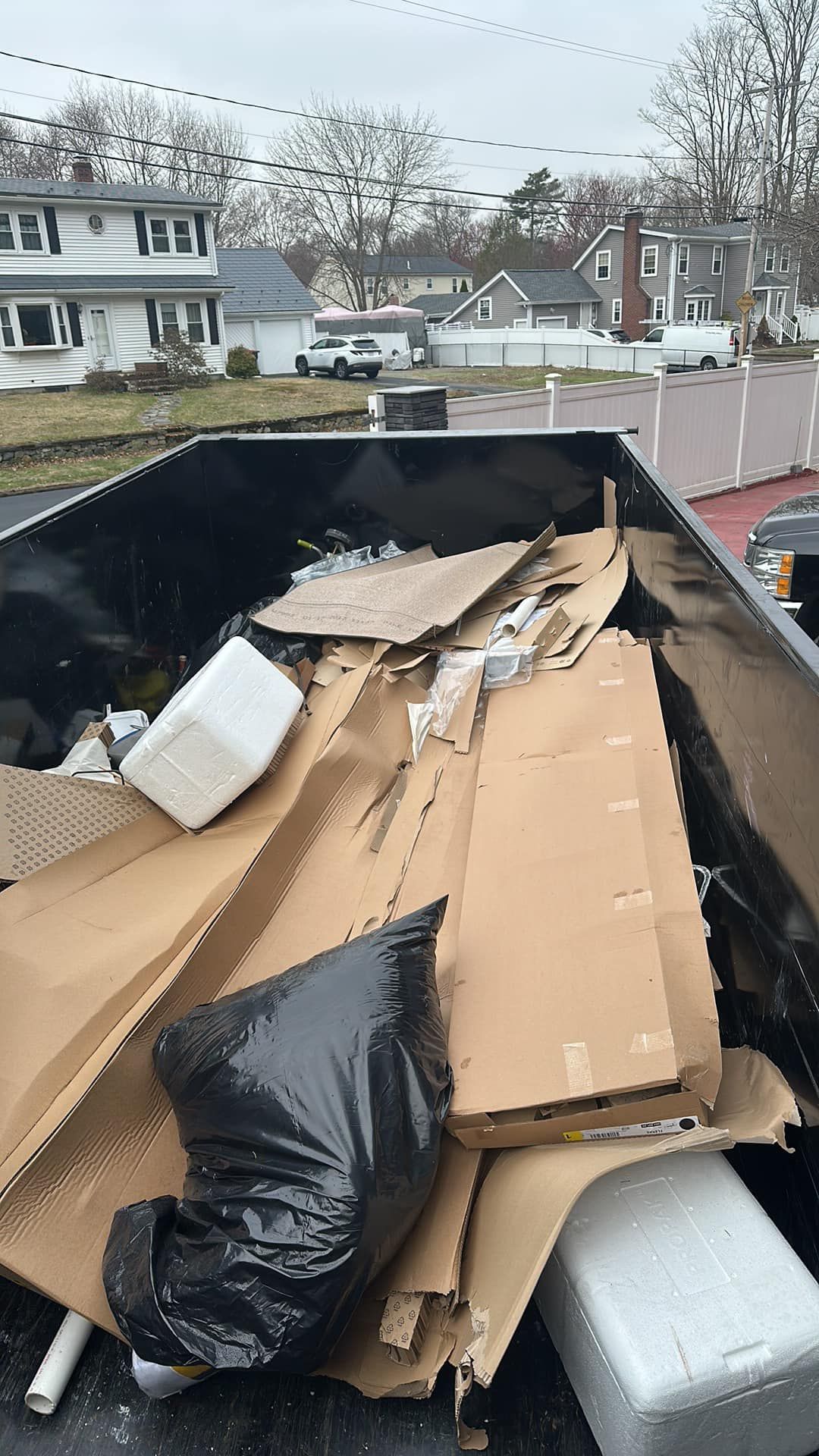 Dumpster filled with cardboard boxes, styrofoam, and a black trash bag, outdoors in a neighborhood.