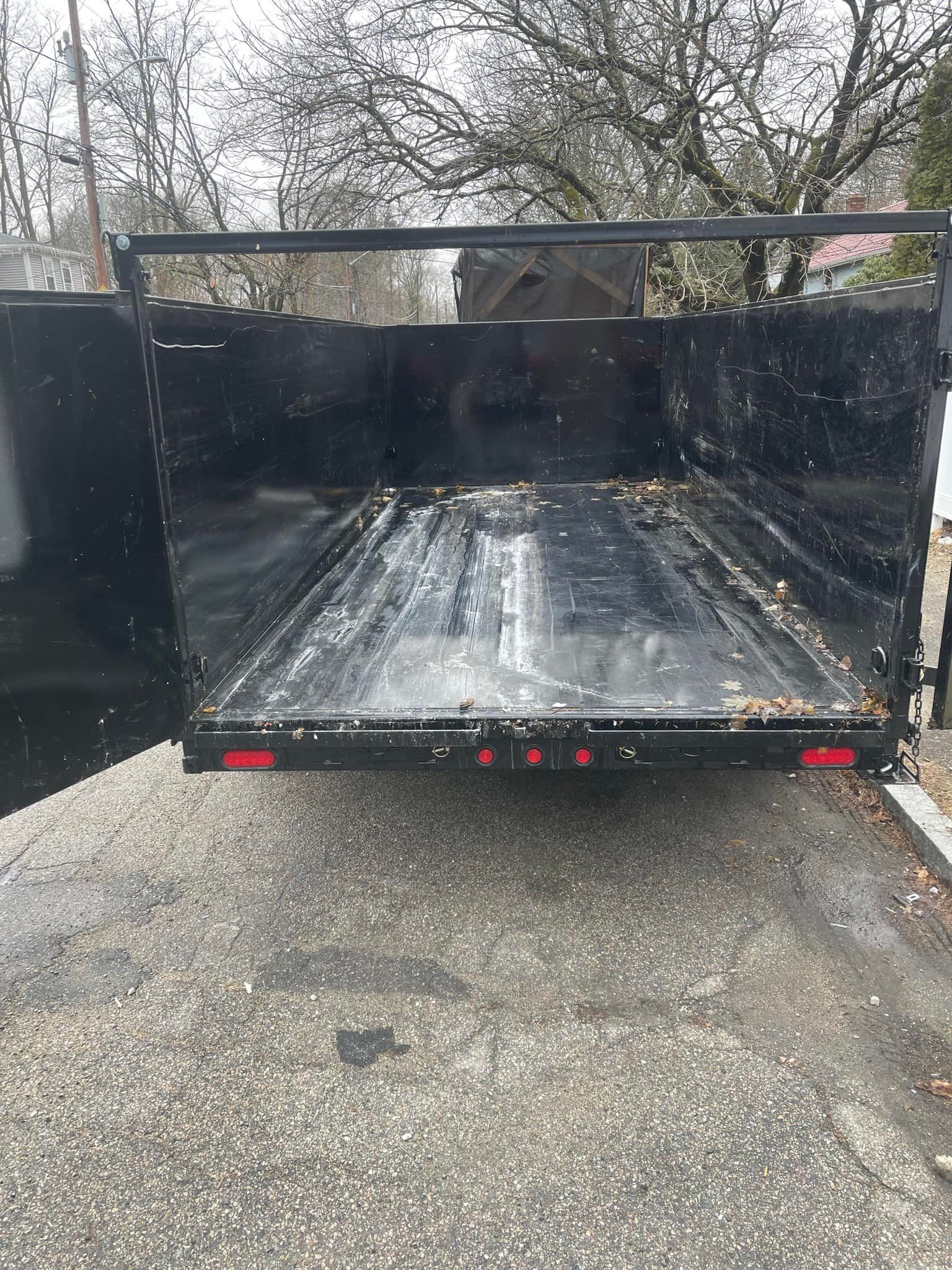 Black trailer bed, open, facing away. Tail lights visible. Gravel ground.
