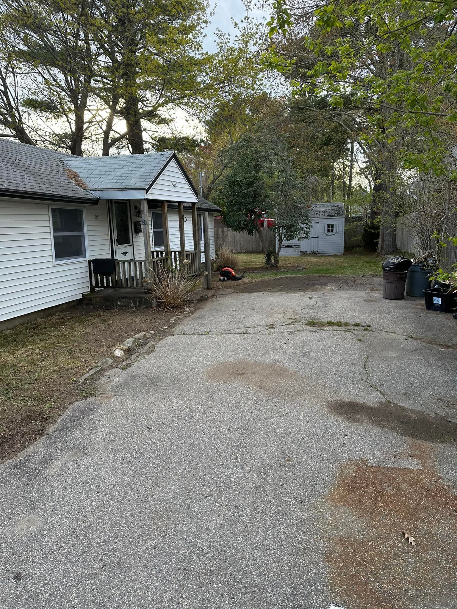 Gravel driveway leads to a white house with a small porch, surrounded by trees and a shed.