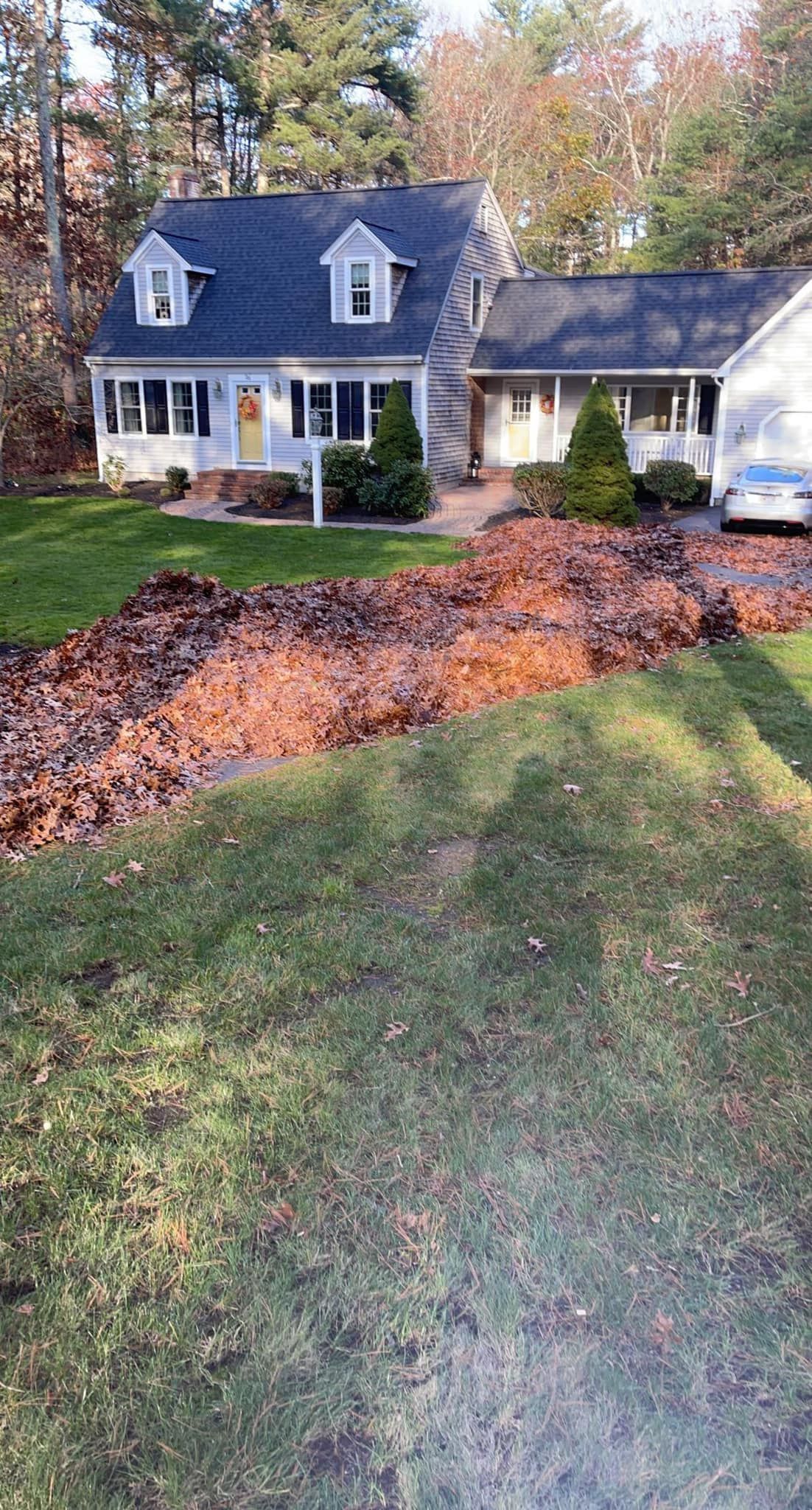 House with blue roof, dormers, and white trim, fronted by a large pile of fallen leaves, on a grassy lawn.