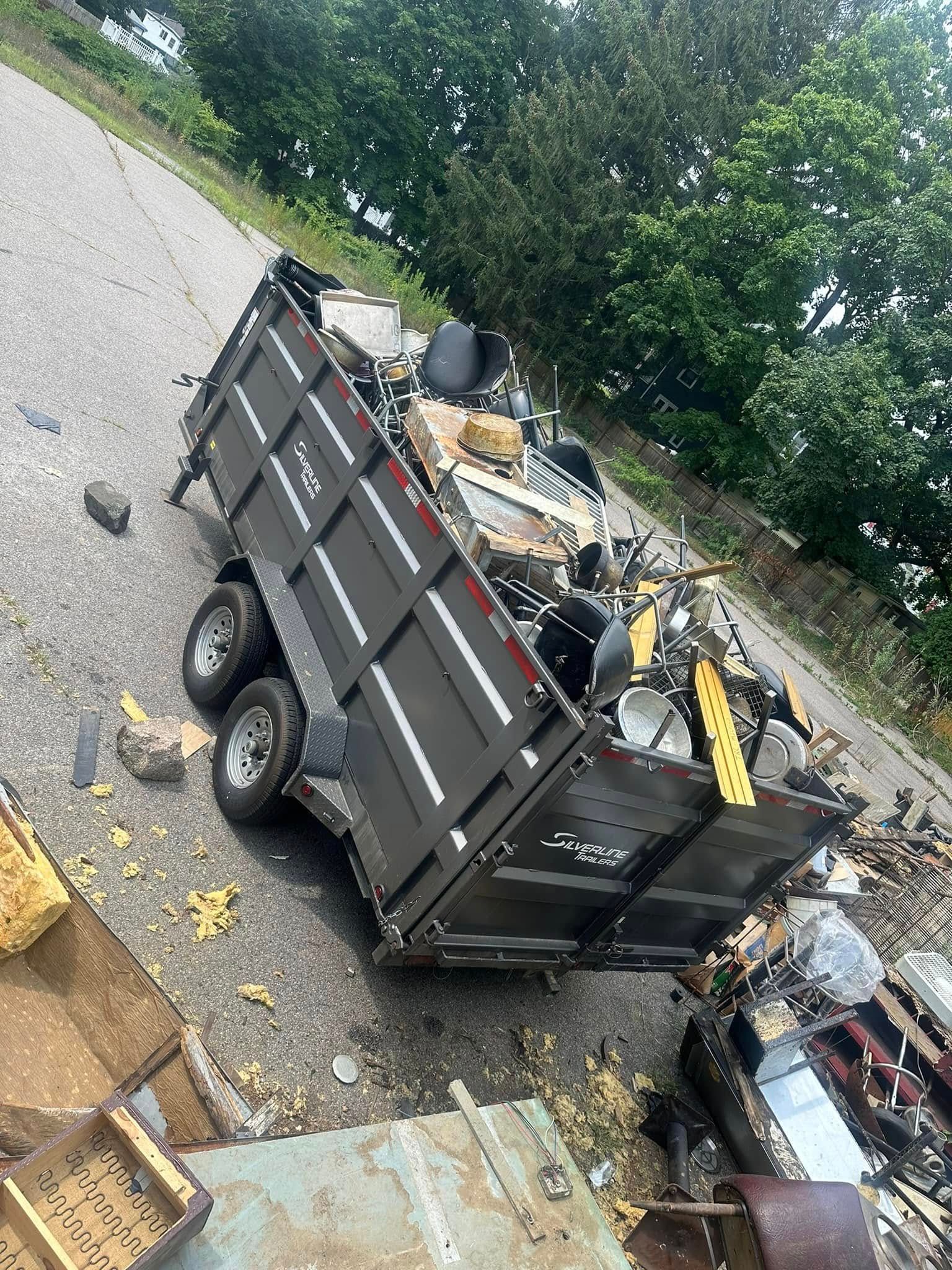 A loaded black trailer filled with debris sits on a gravel surface next to a metal bin.