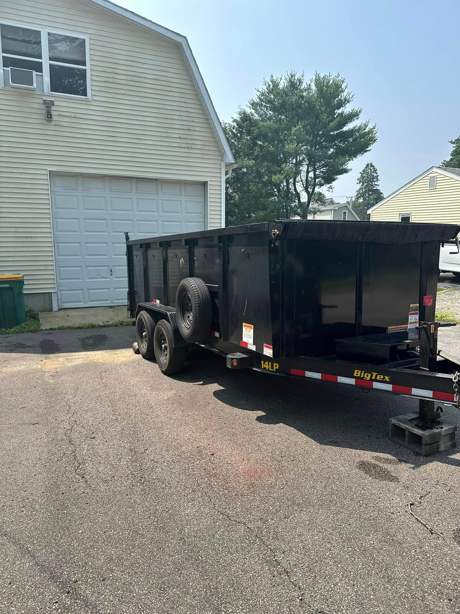 Black dump trailer parked on gravel, in front of a white garage.