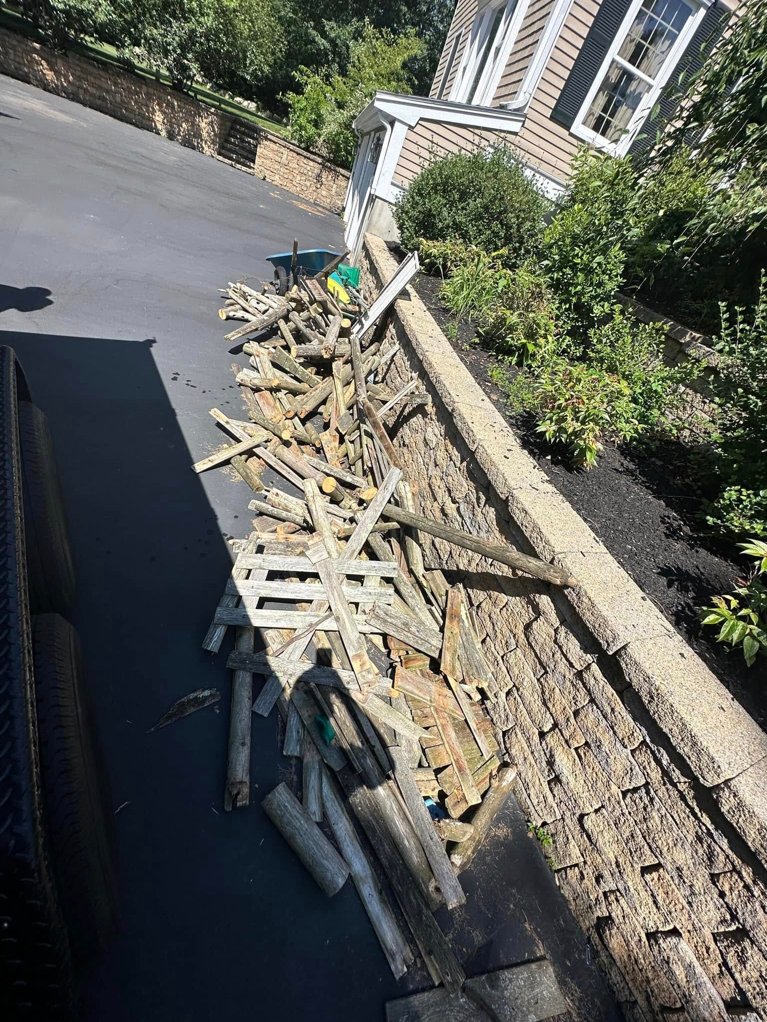 Pile of broken wood debris next to a retaining wall and black asphalt driveway. House in background.