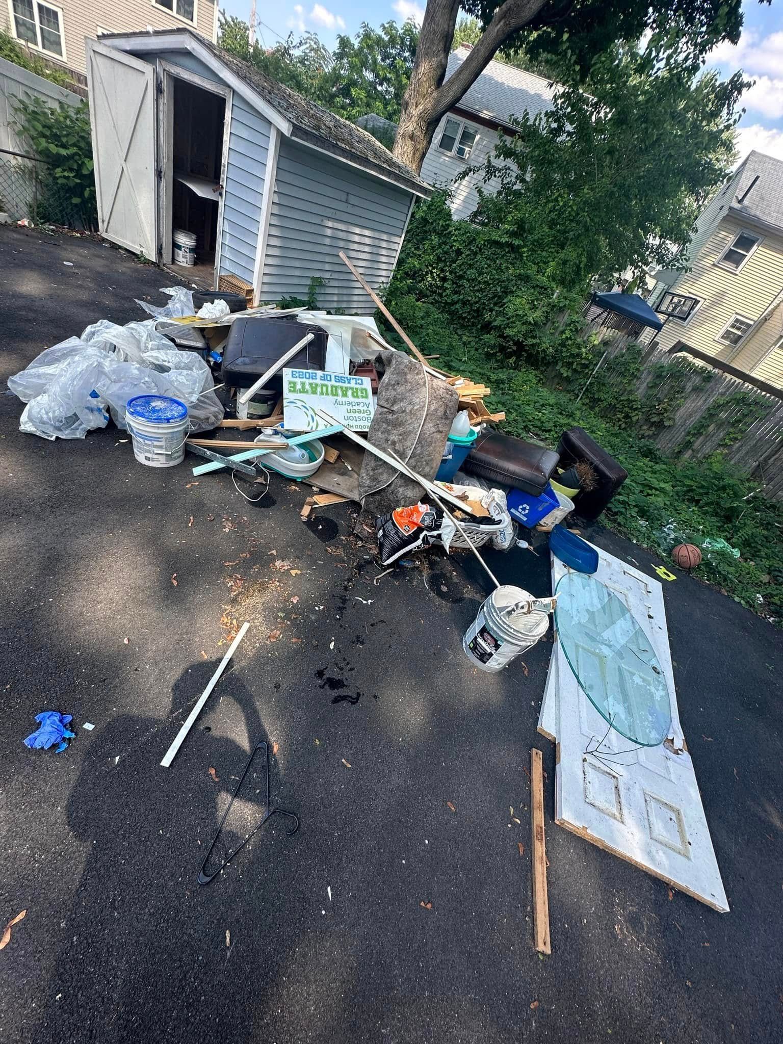 Asphalt driveway littered with trash; a light blue shed sits in the background.