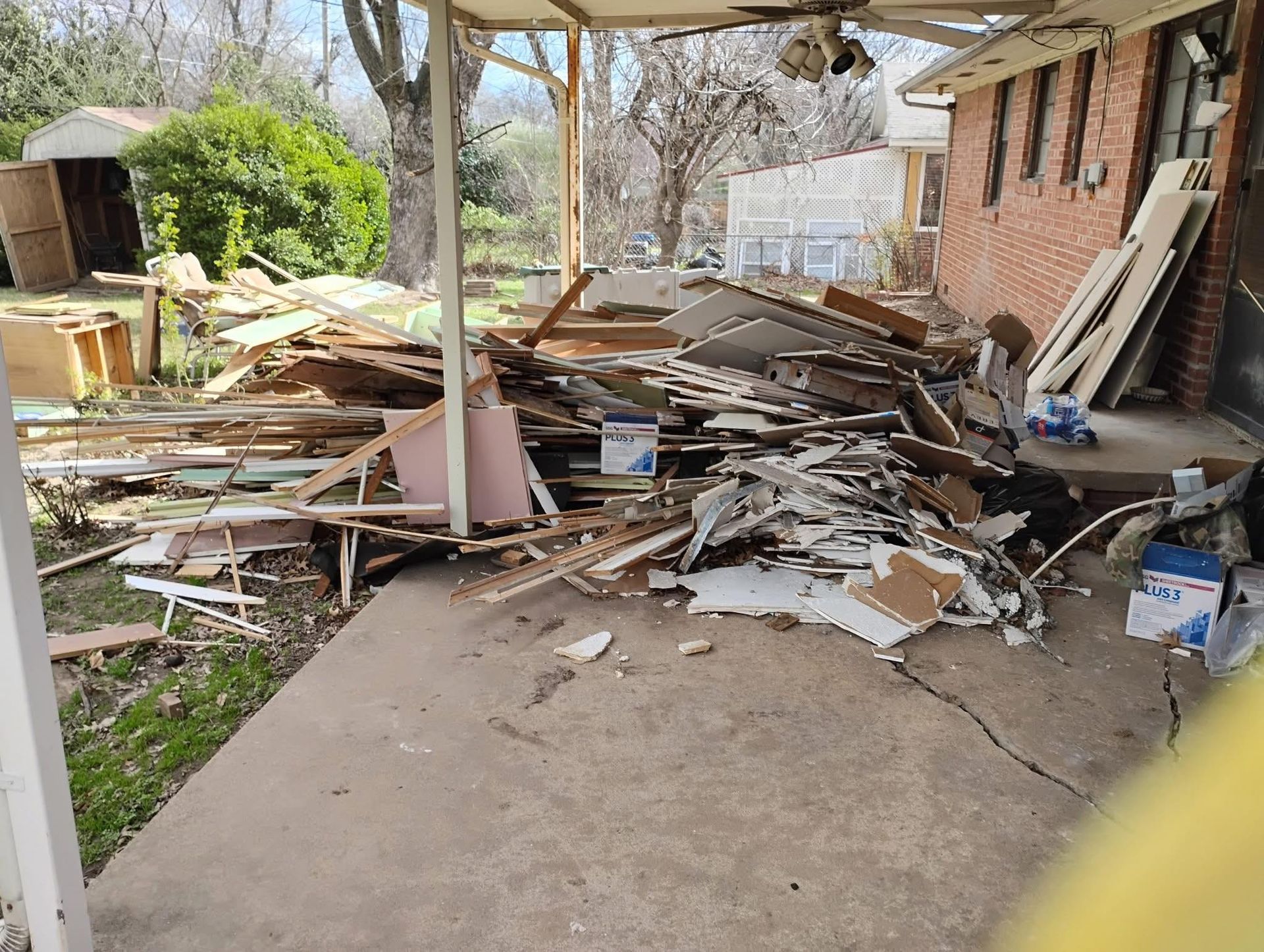 Exterior view of a house with debris pile on the porch. Brown bricks, concrete floor, and a weathered wood fence.