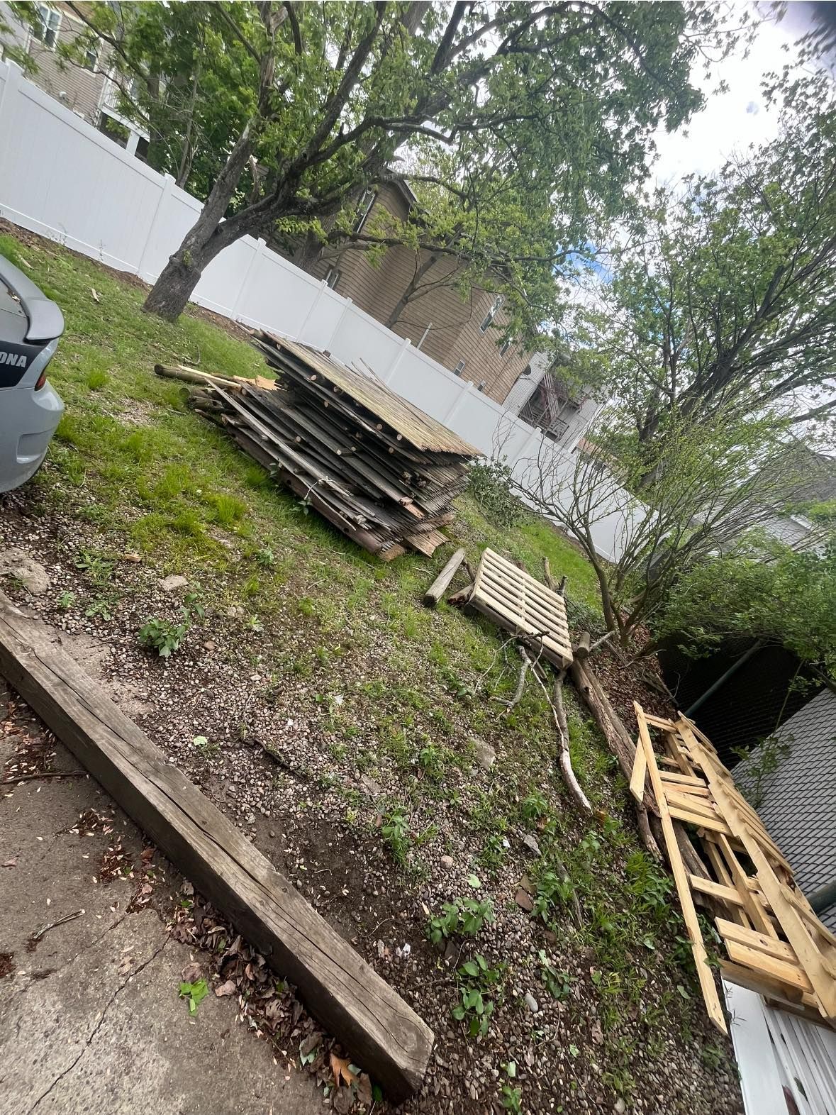 Pile of wood and debris on a grassy, sloped yard next to a white fence.