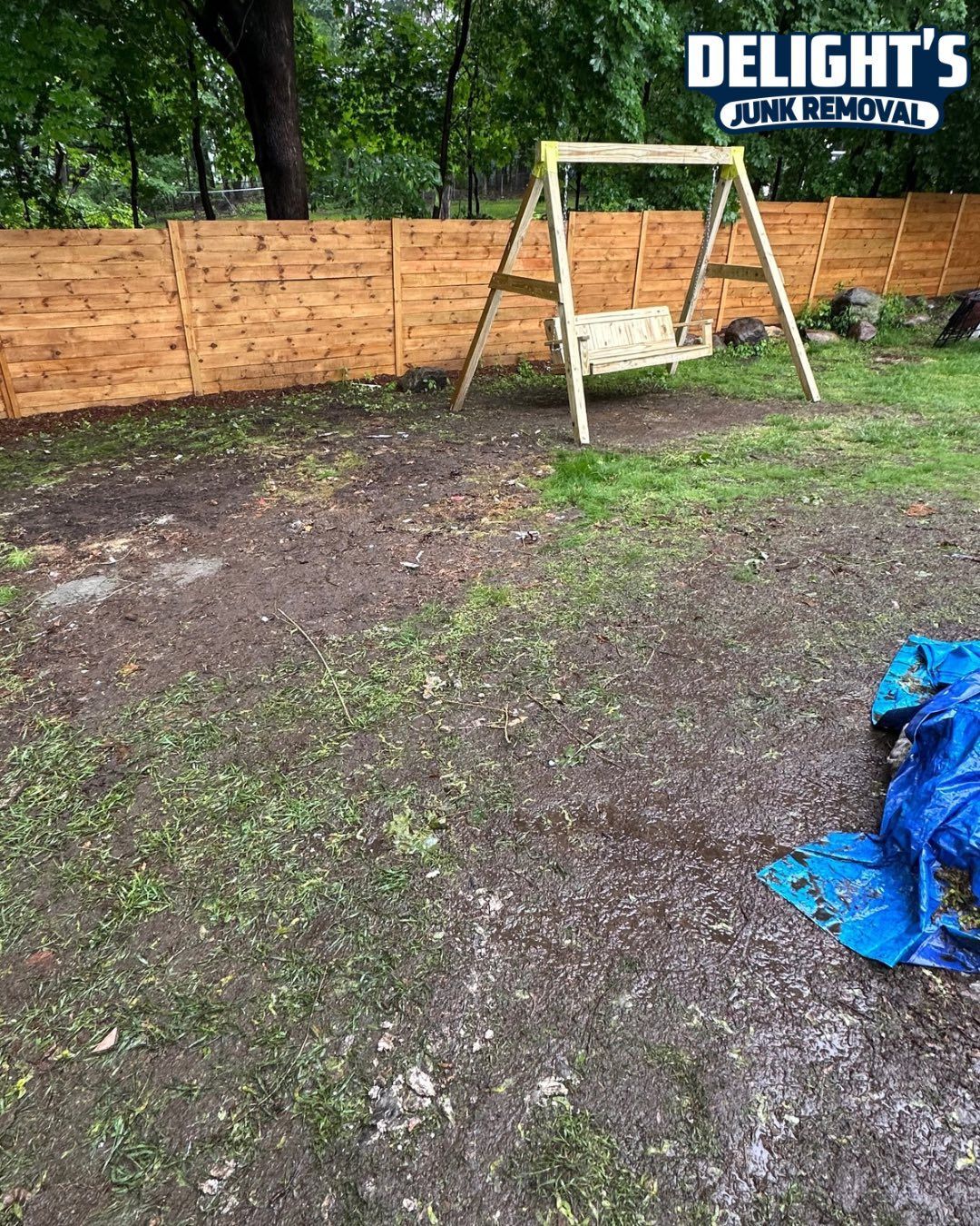 Backyard with a swing set and wooden fence. The ground is muddy, and there is a blue tarp.