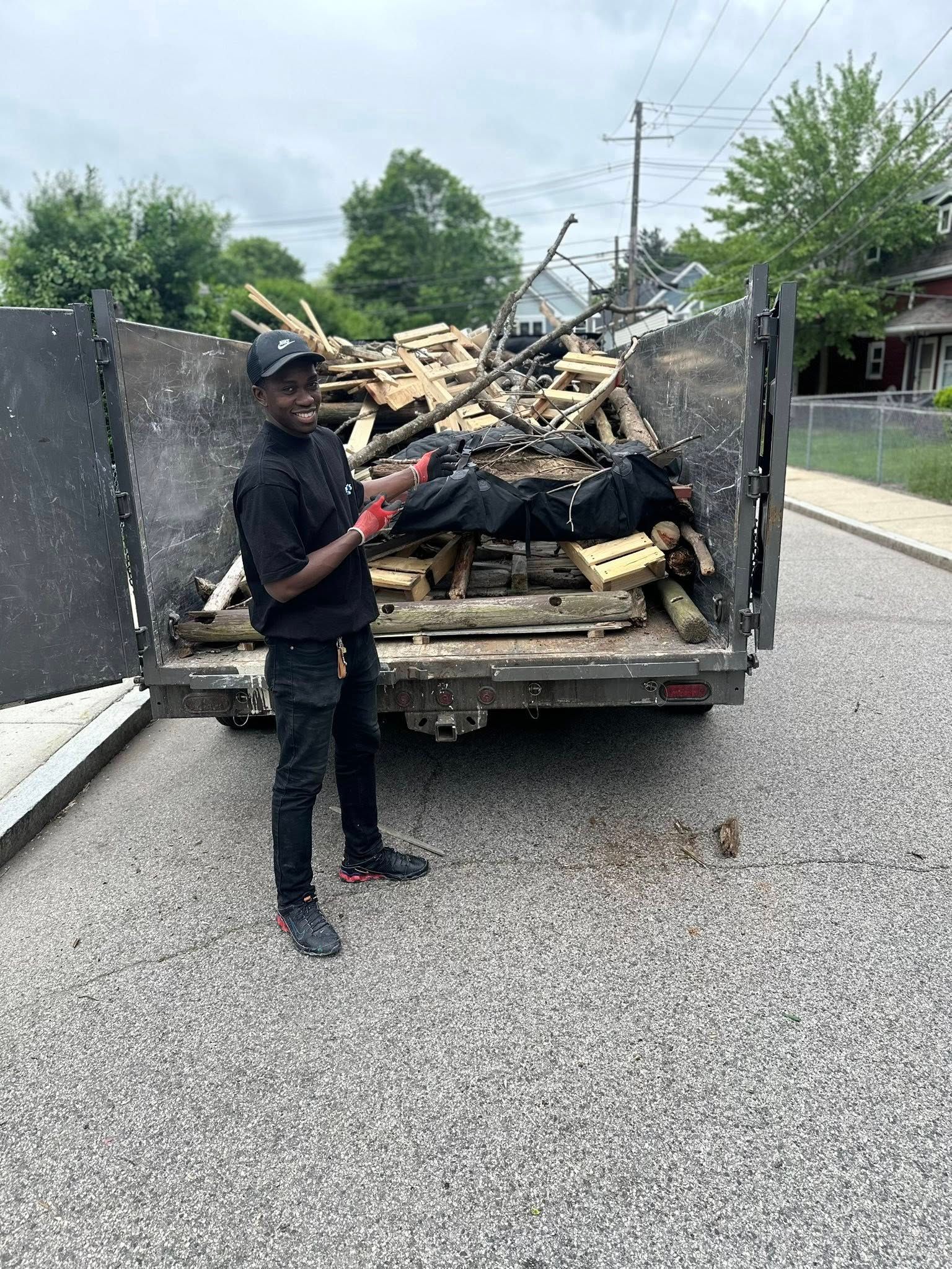 Man standing in front of a full truckload of wood, pointing, wearing black clothing, outdoors on pavement.