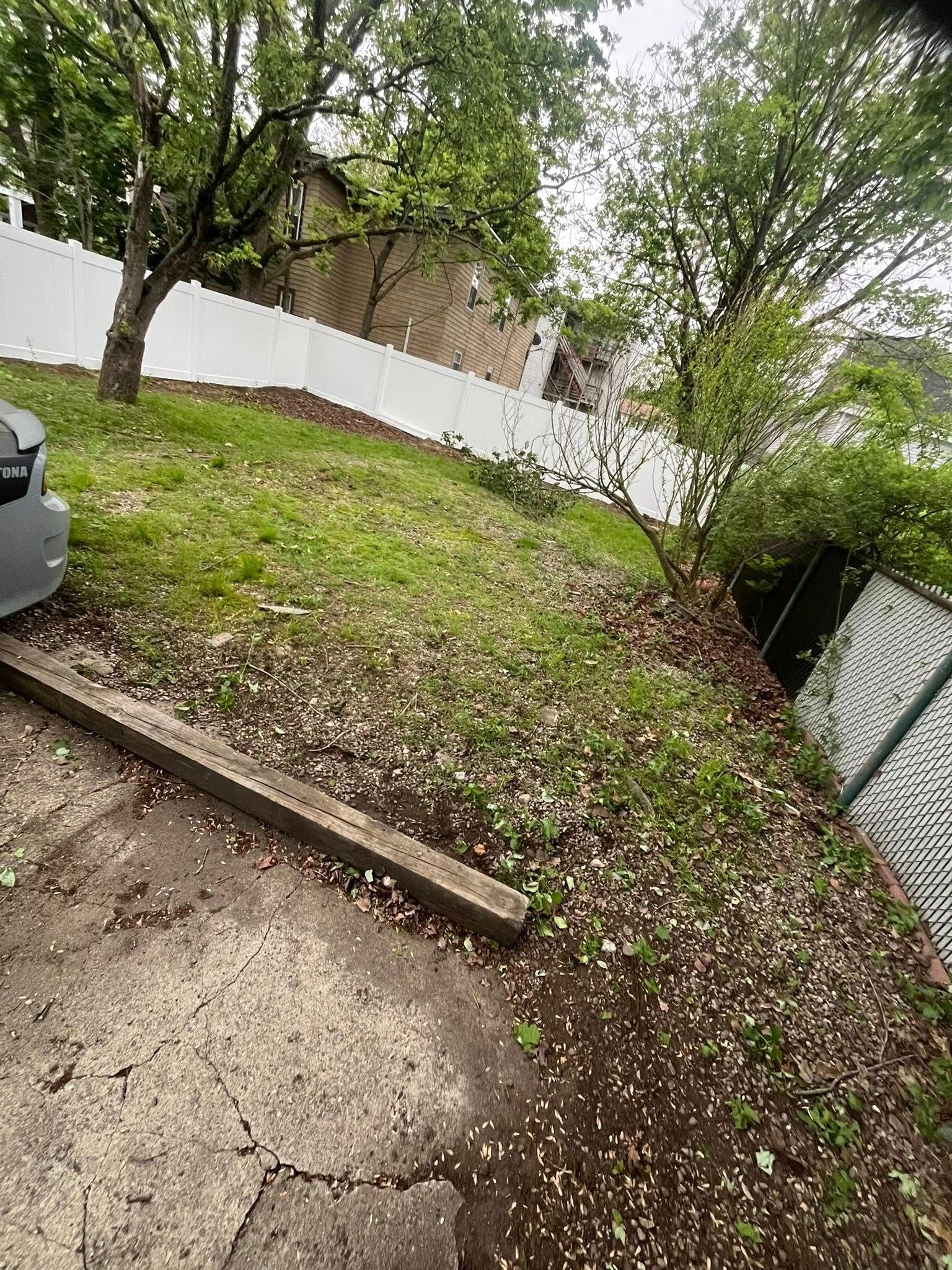 Sloped, grassy yard with a white fence.  A gray car, trees and a wooden step are visible.