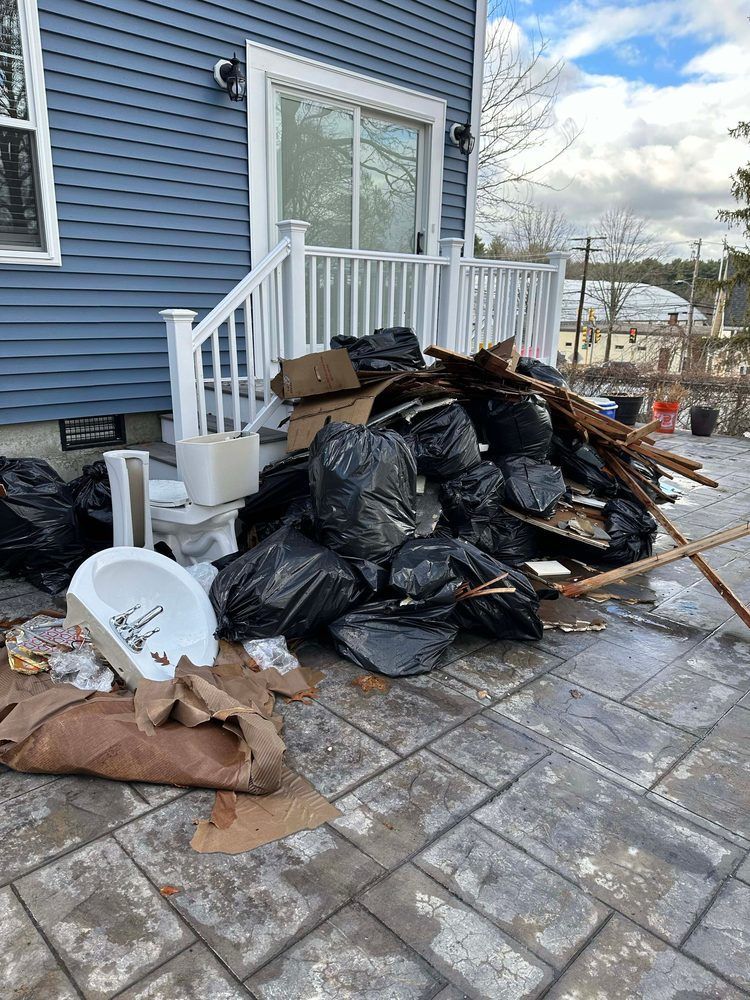 Pile of black trash bags, debris, and a toilet on a patio beside a blue house with a white railing.