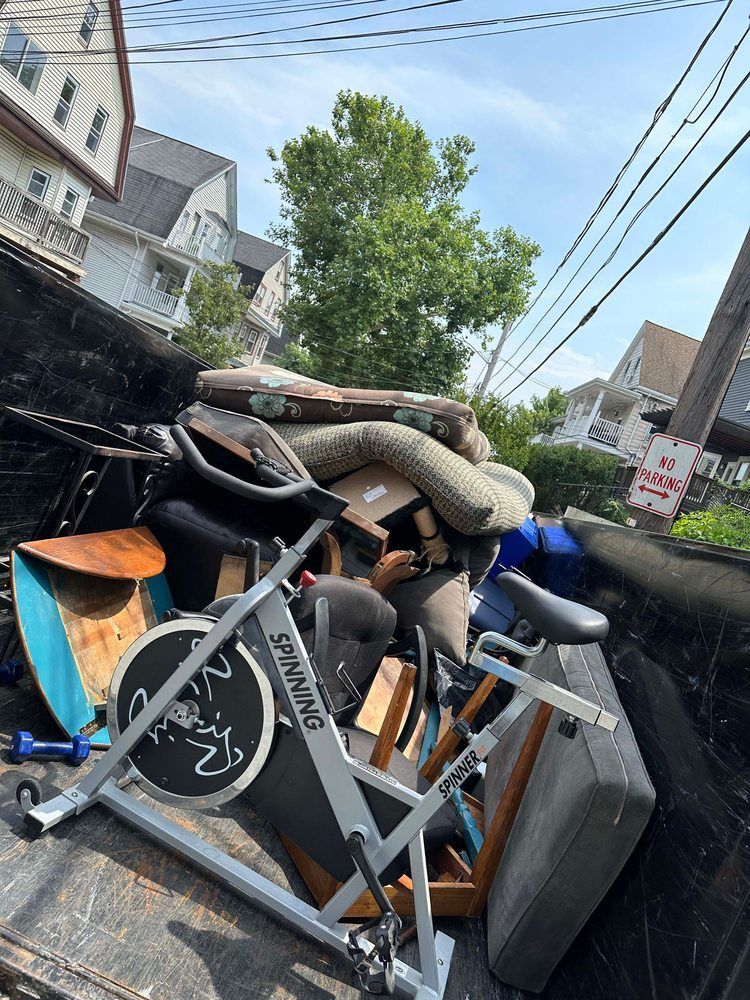 A dumpster filled with old furniture, including a spin bike, against a backdrop of houses and a tree.