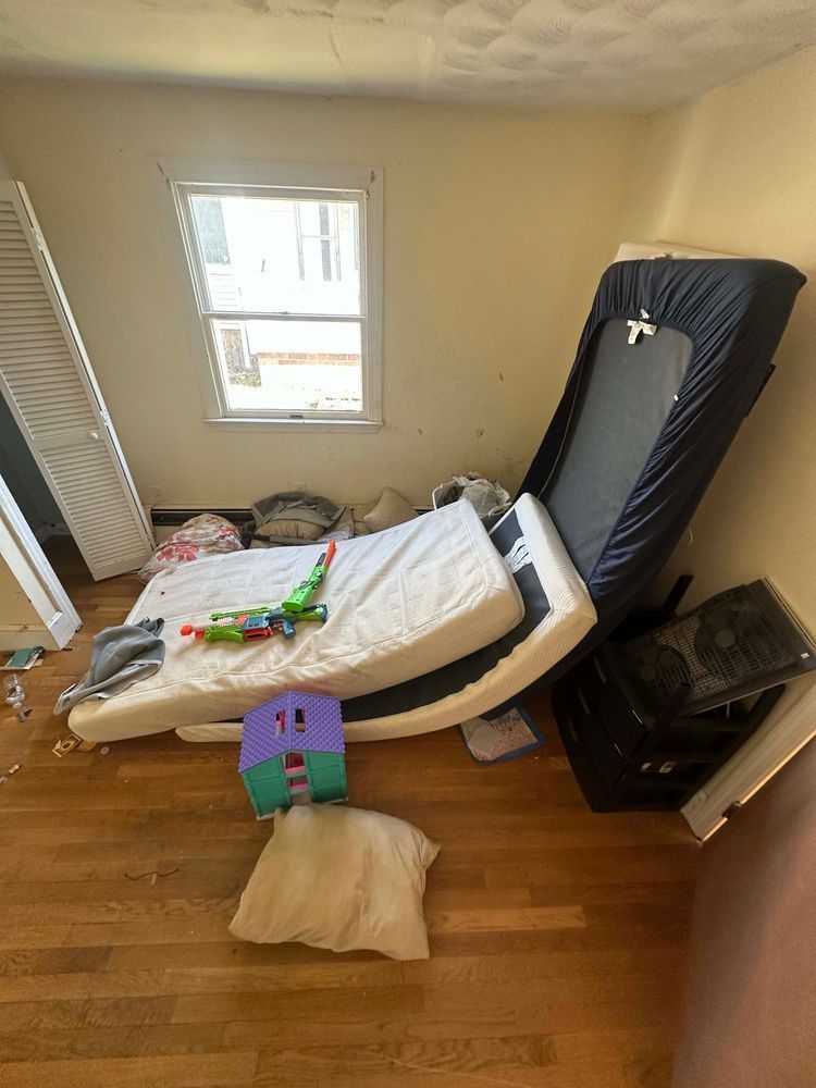 Room with scattered mattresses, toys, and debris. Includes a small window and wooden floors.