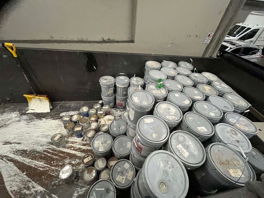 Dumpster filled with paint cans; some open, spillage visible, shovel nearby.