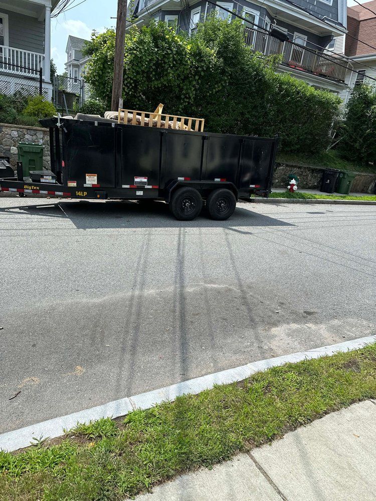 Black trailer with wooden pallets on a street, houses in the background.