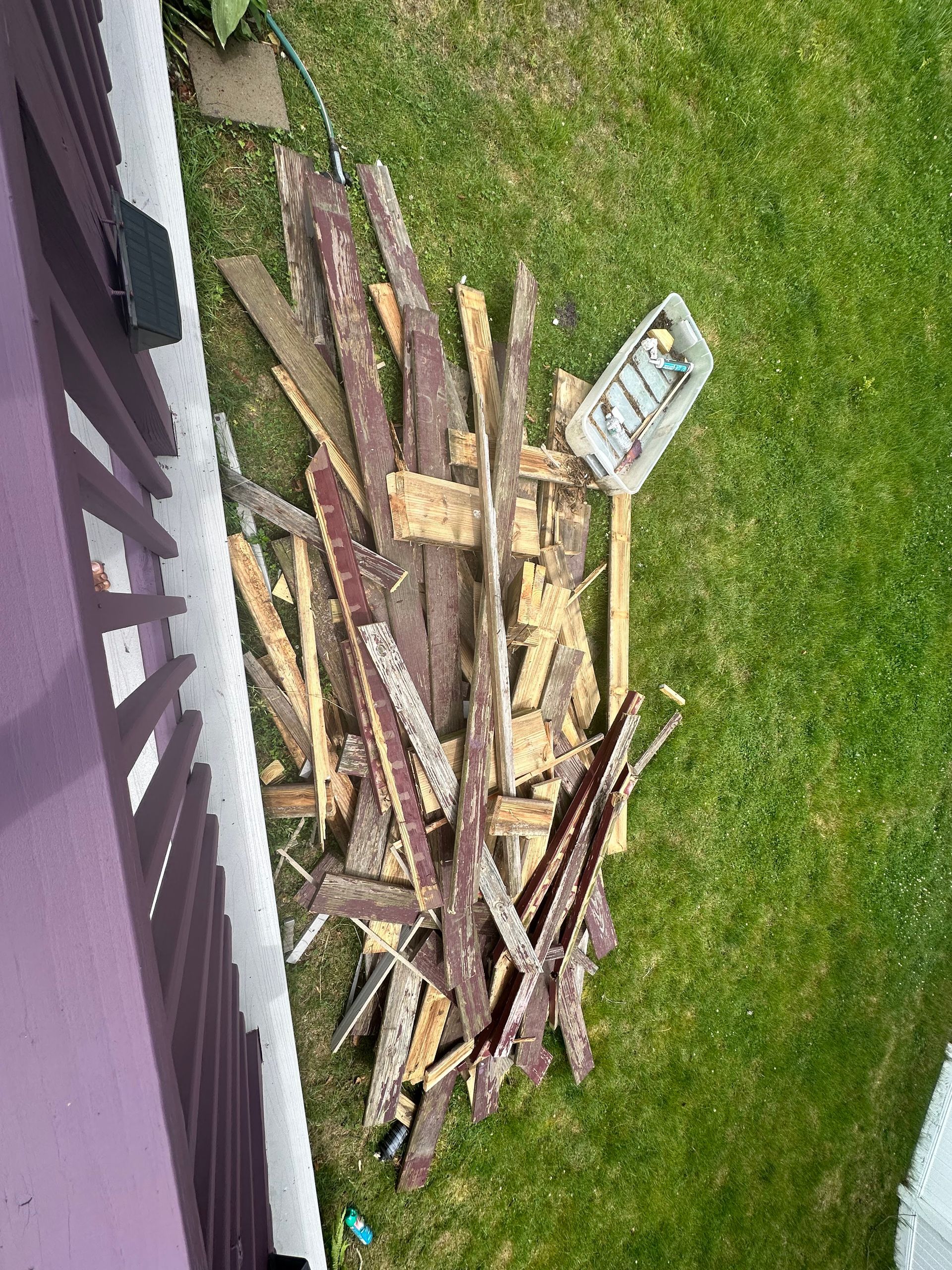 Pile of splintered wood and debris on grass next to a purple porch railing.