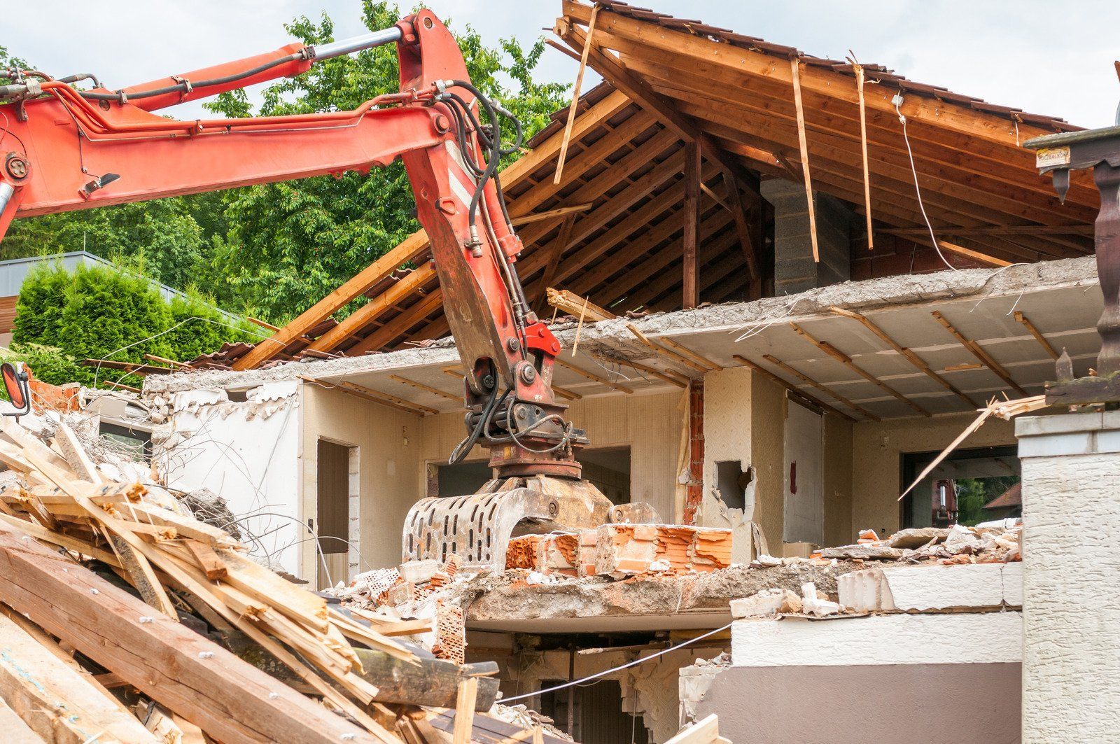 An excavator demolishes a house, debris and the roof visible.