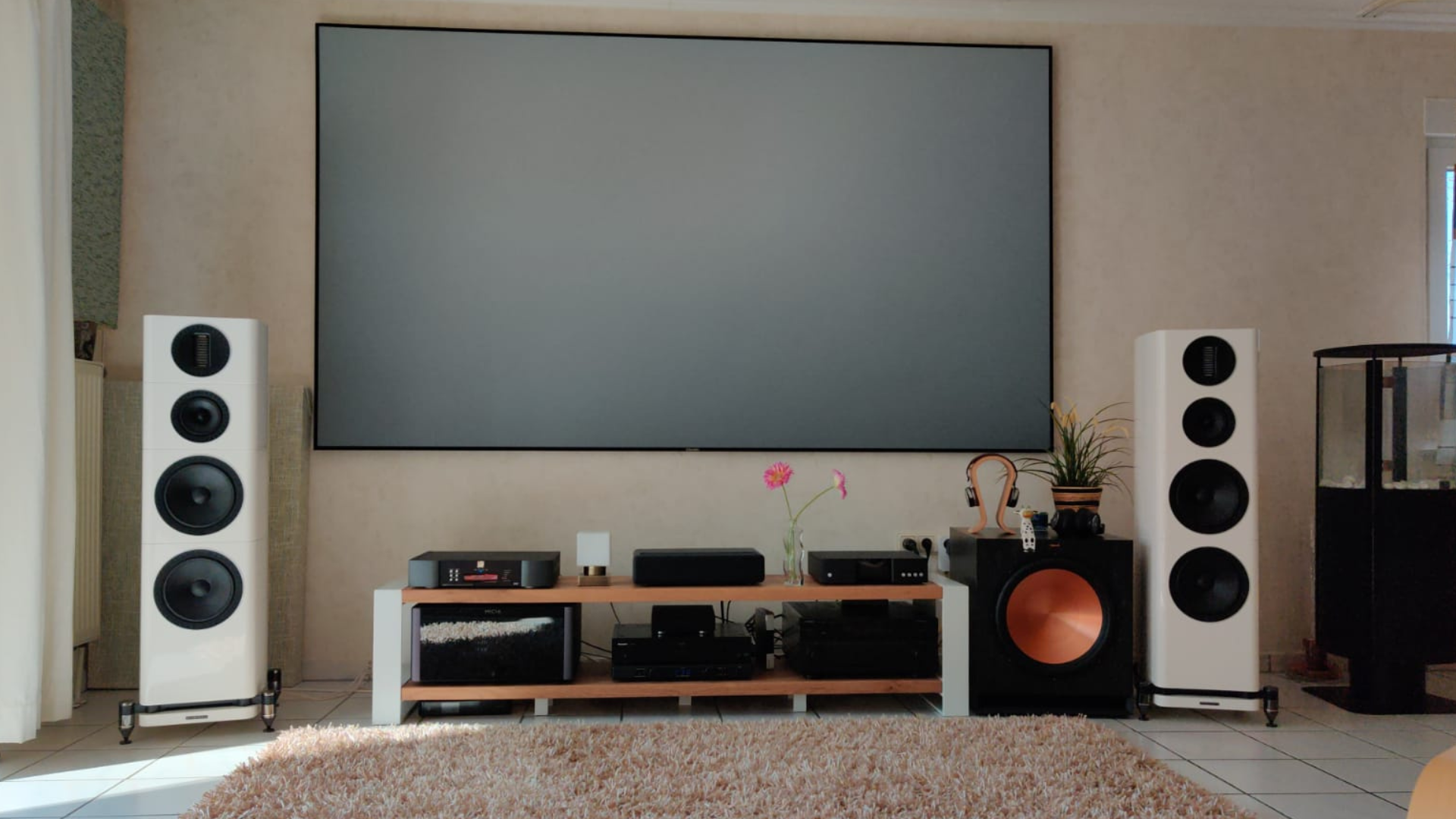 Home theater setup: white speakers flank a large screen, audio components sit on a wooden console.