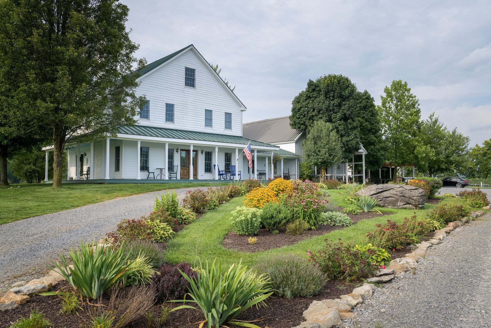 A white house with a large porch is surrounded by flowers and trees.