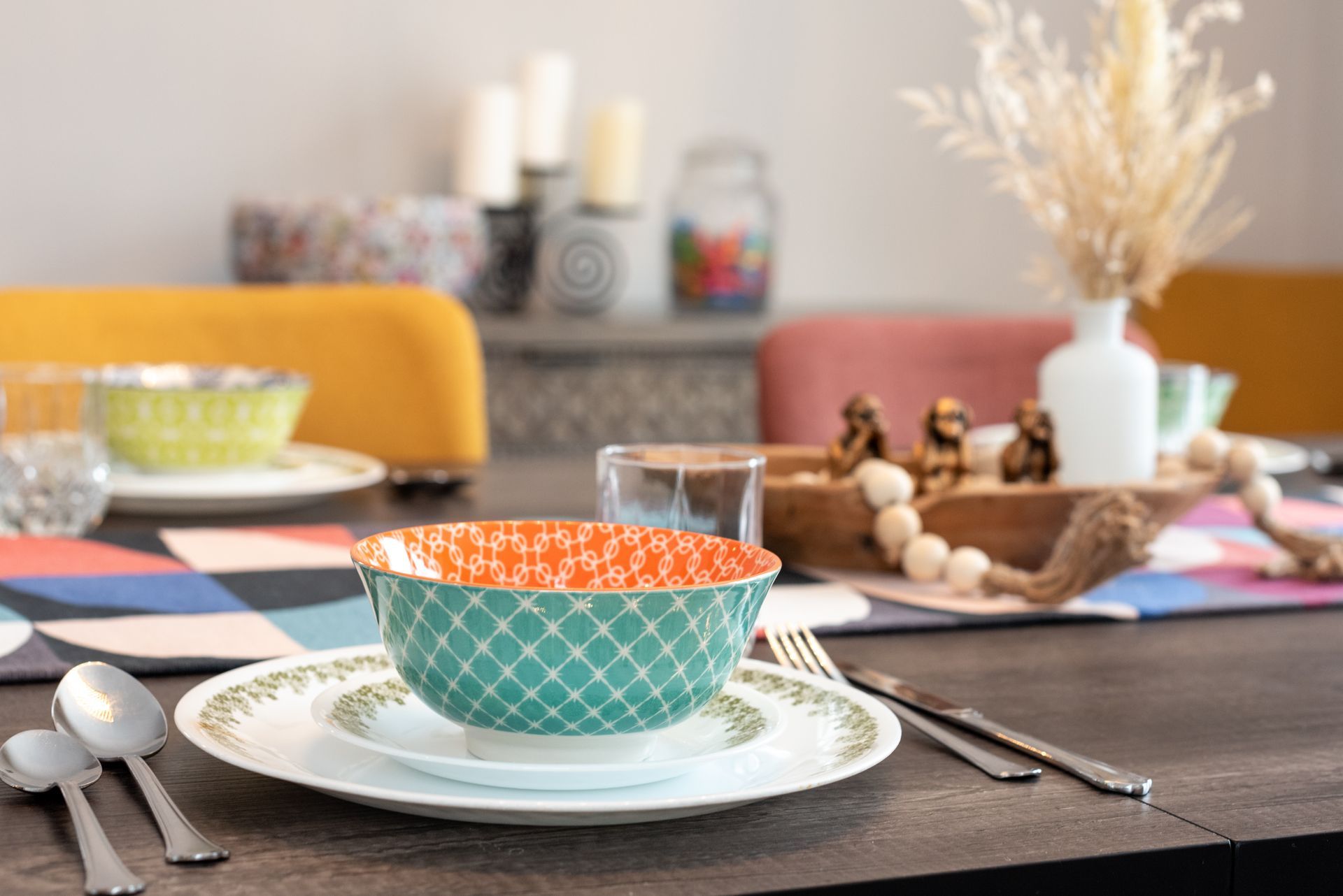 A dining room table with plates , bowls , silverware and a vase of dried flowers.