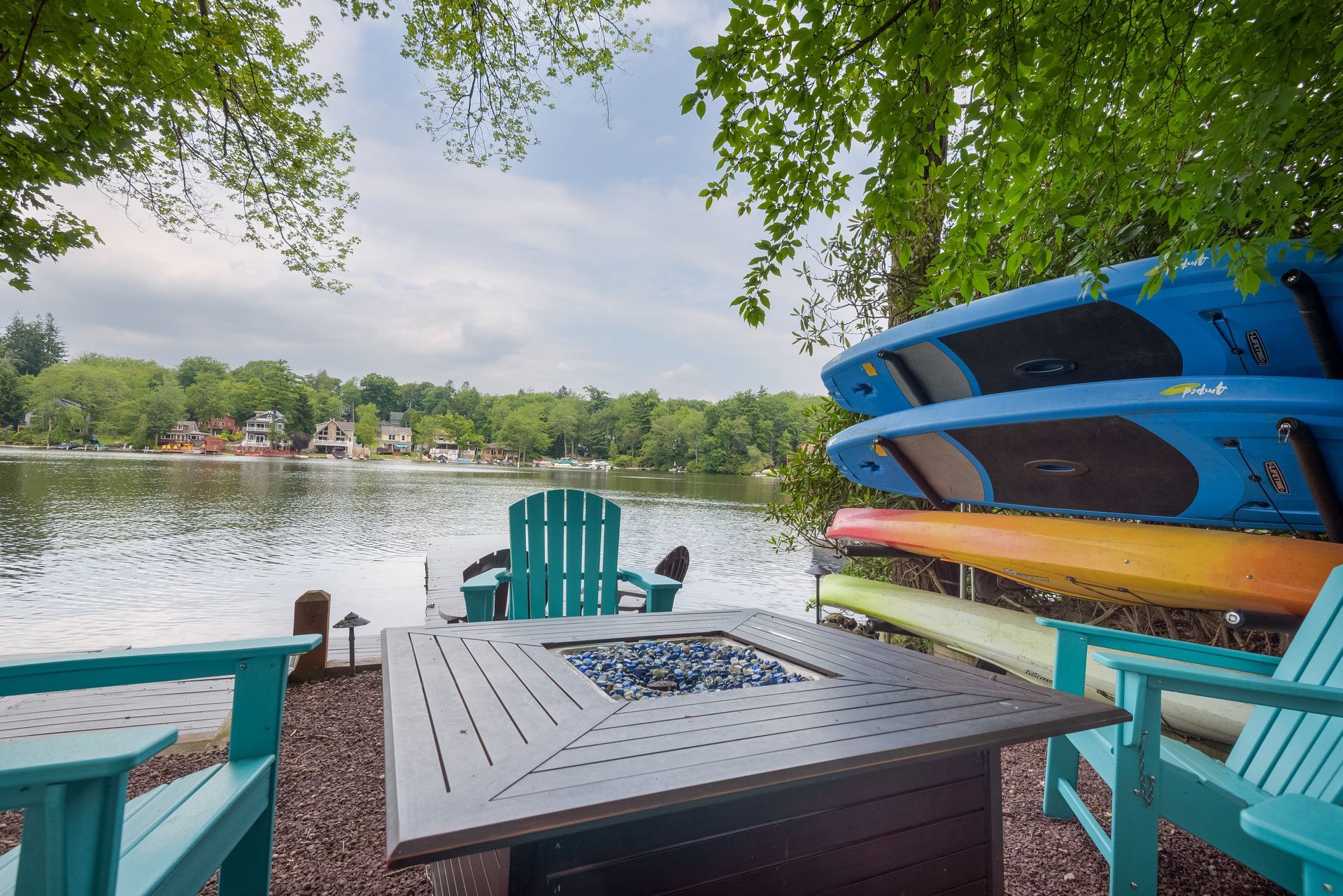A fire pit is sitting next to a lake surrounded by chairs and kayaks.