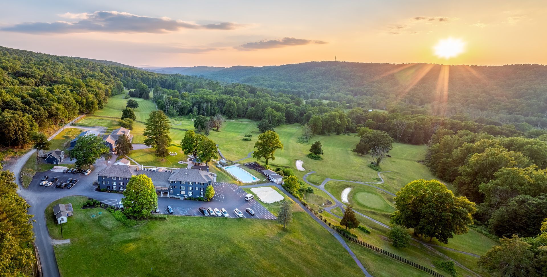 An aerial view of a golf course at sunset with the sun shining through the clouds.