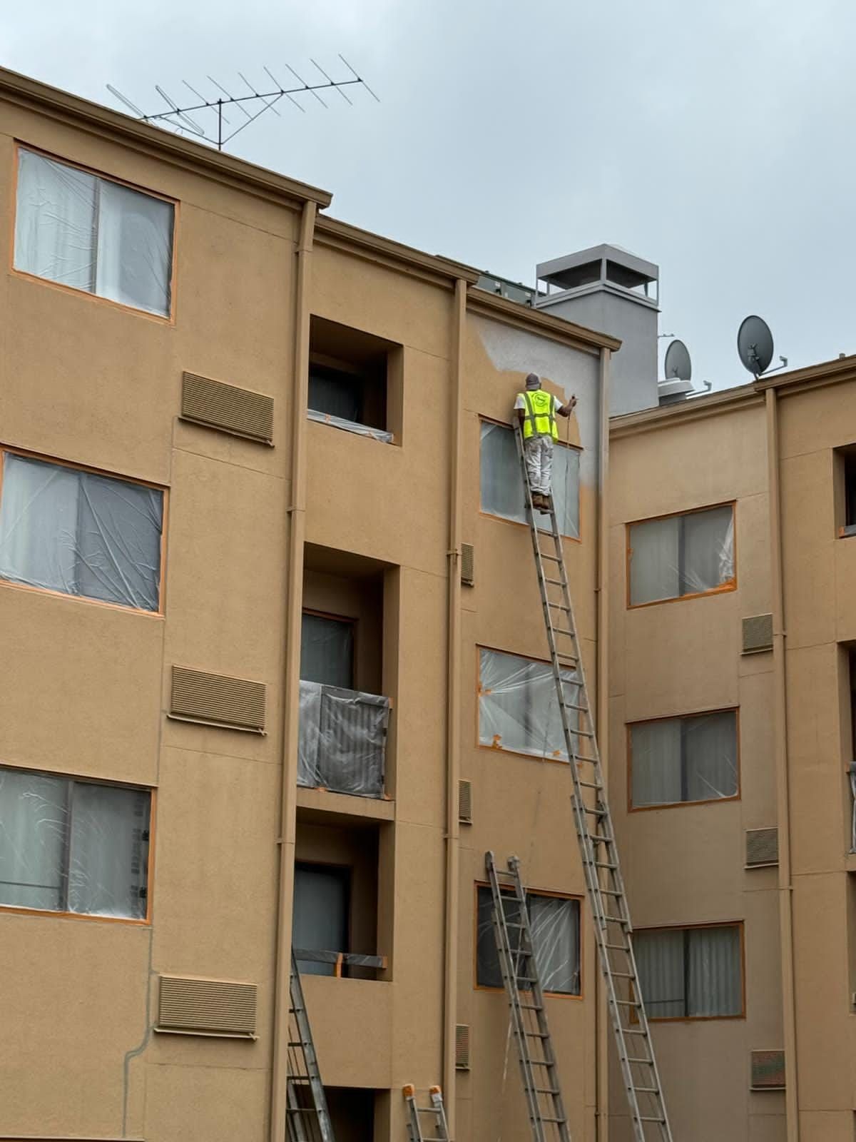 A person on a ladder paints a tan apartment building, wearing a safety vest.