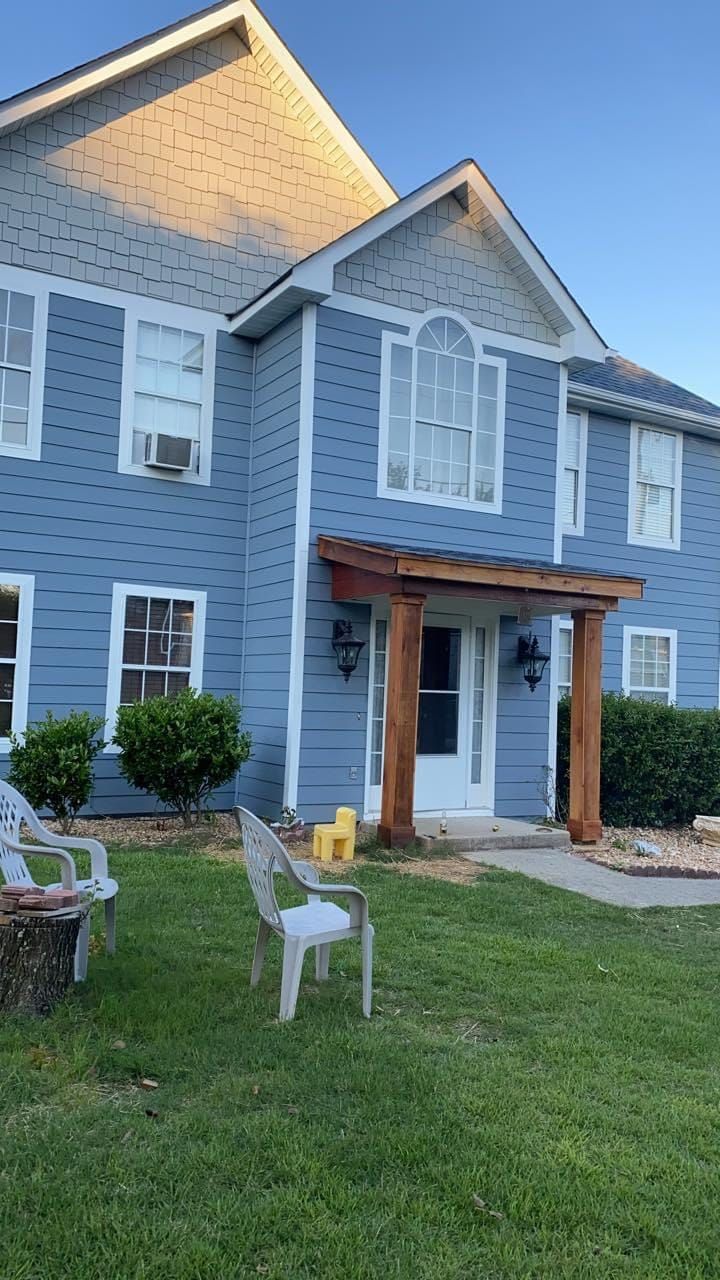 Two-story blue house with wooden porch, chairs on lawn, and shrubs.