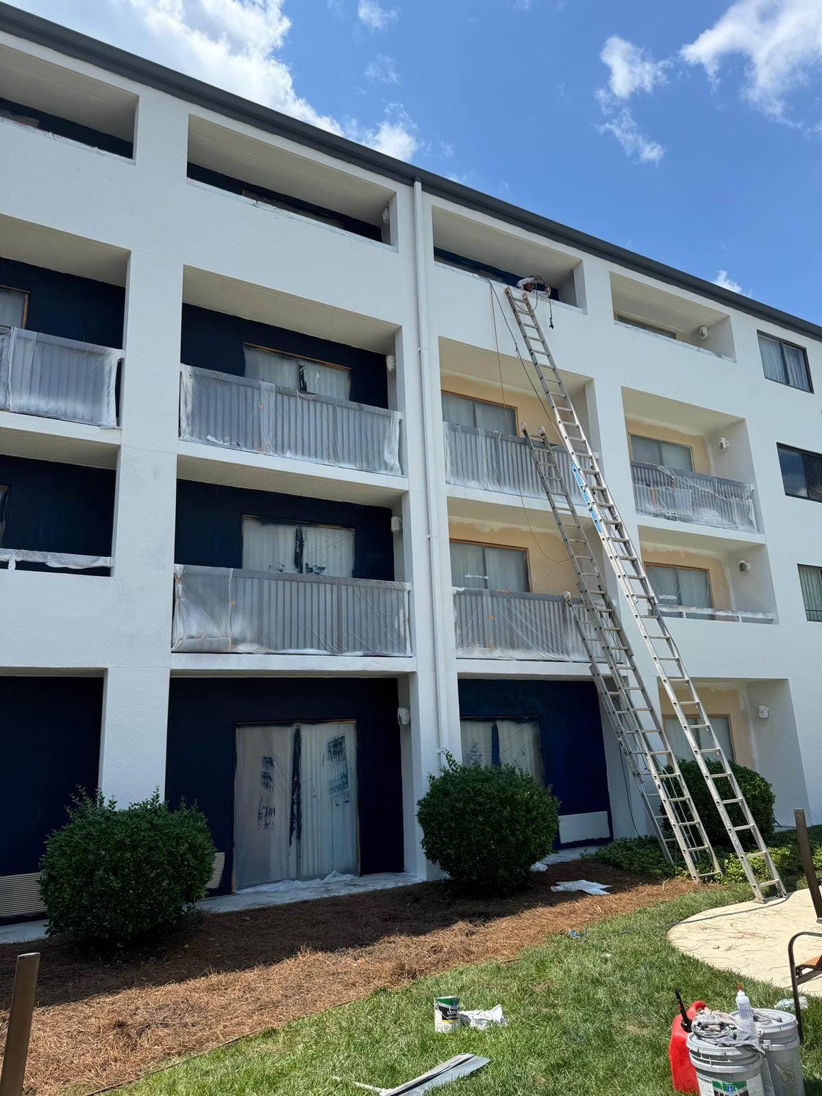 Apartment building exterior being painted. A tall ladder leans against the building. Sky is blue with clouds.