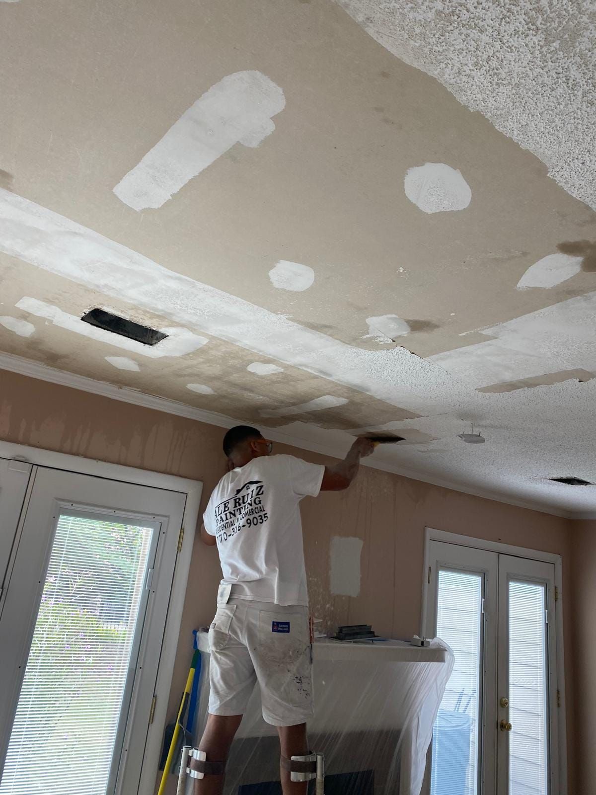 Person on a ladder repairs ceiling with patching compound. Interior shot with windows and a fireplace.