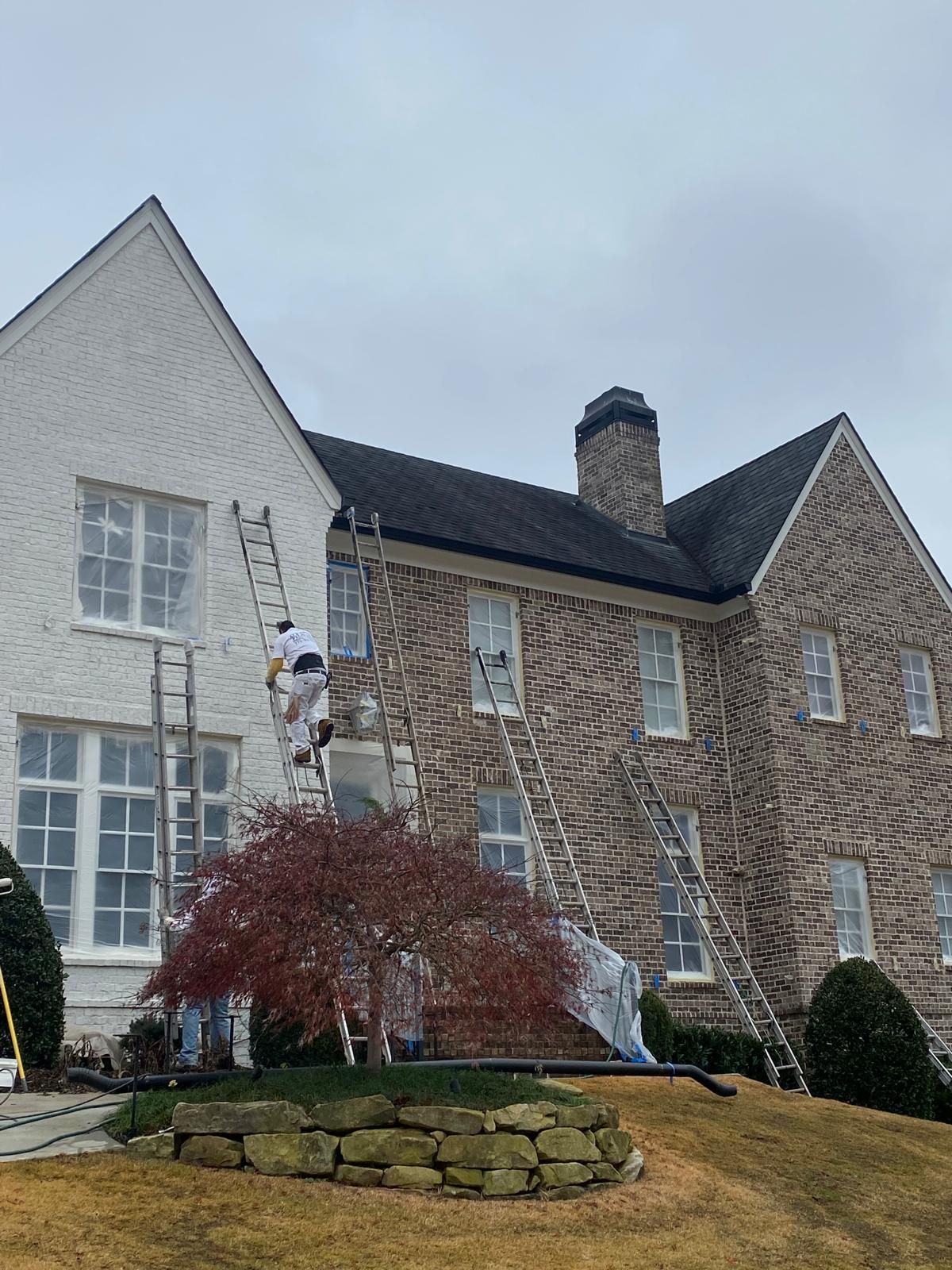 Workers on ladders painting the exterior of a two-story house with stone and white siding. Overcast sky.