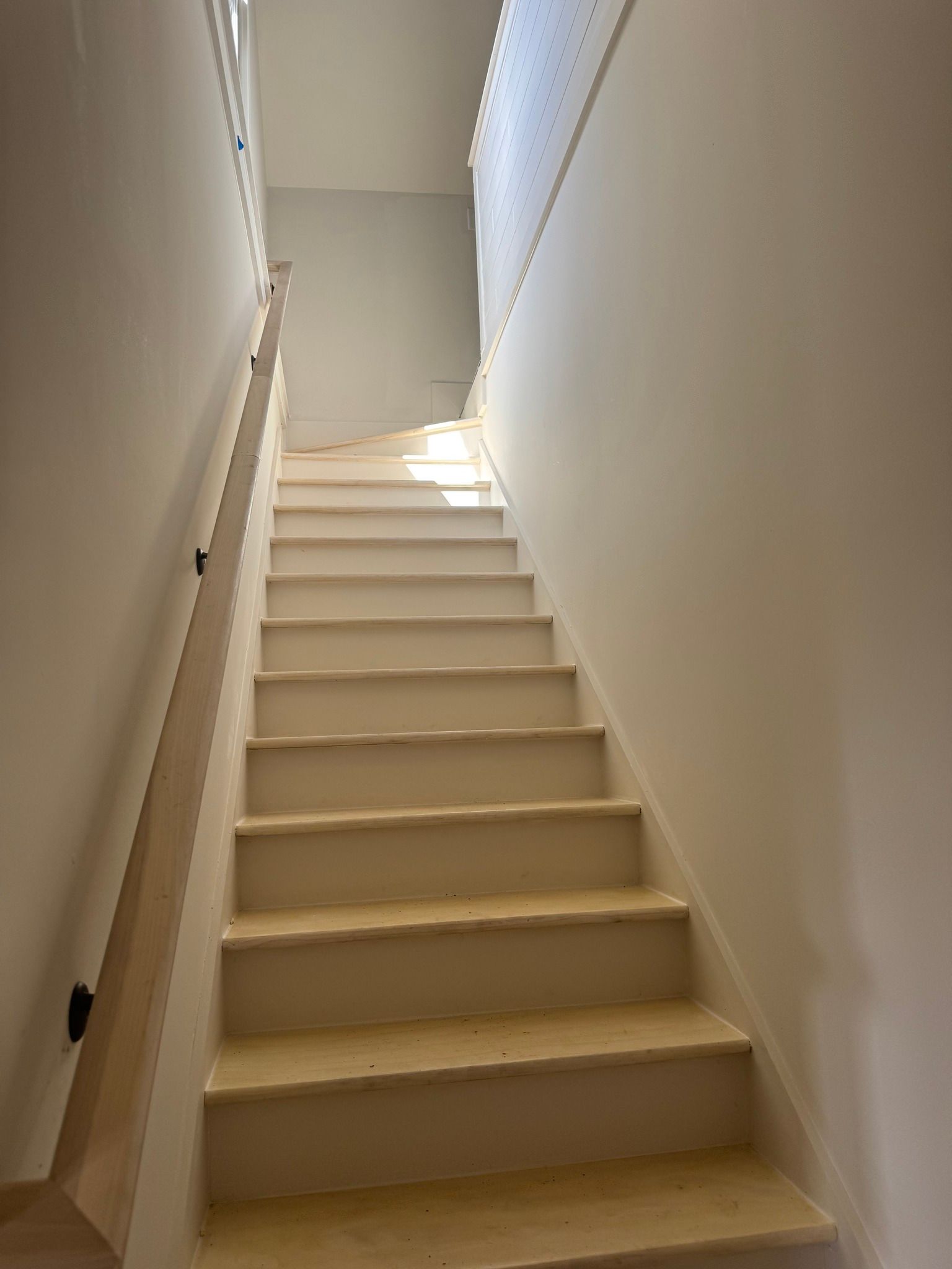 Staircase with light-colored steps and walls, wooden handrail on the left. Sunlight at the top.