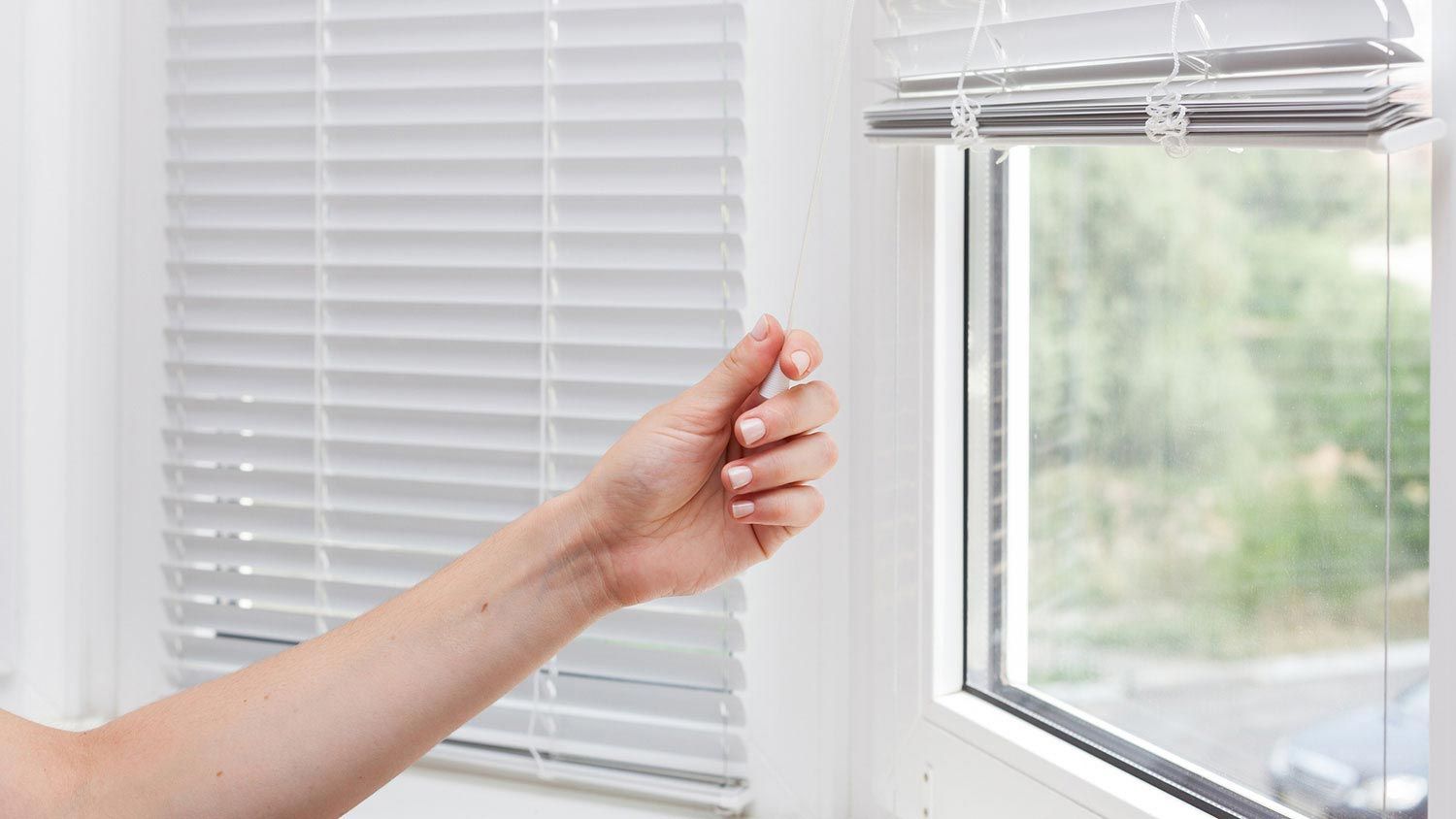 A woman is opening a window with blinds on it.