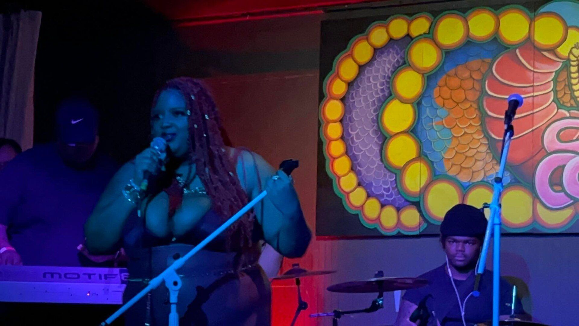 A woman is singing into a microphone on a stage while a man plays drums.