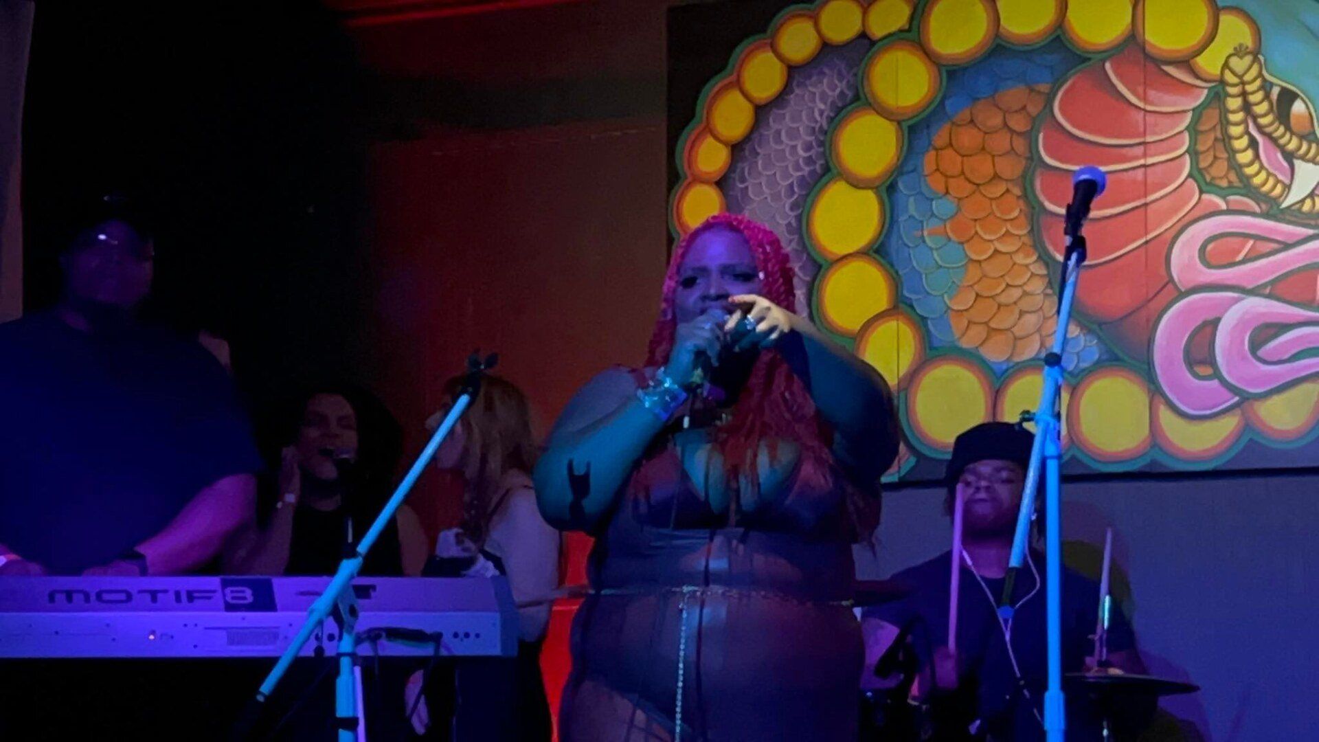 A woman is singing into a microphone on a stage while a man plays drums.