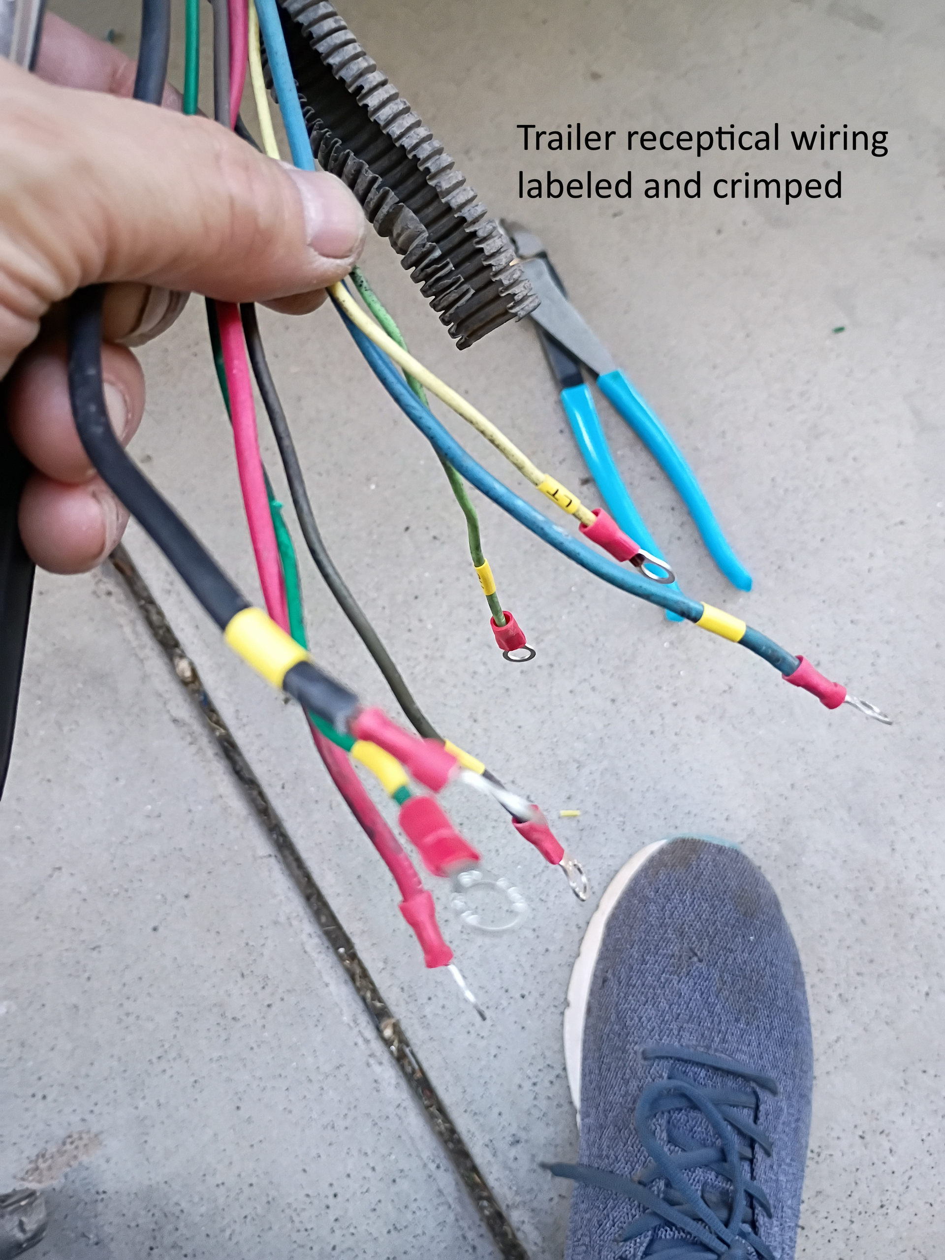 Hand holding labeled trailer receptacle wiring with colored wires and crimped connectors on concrete