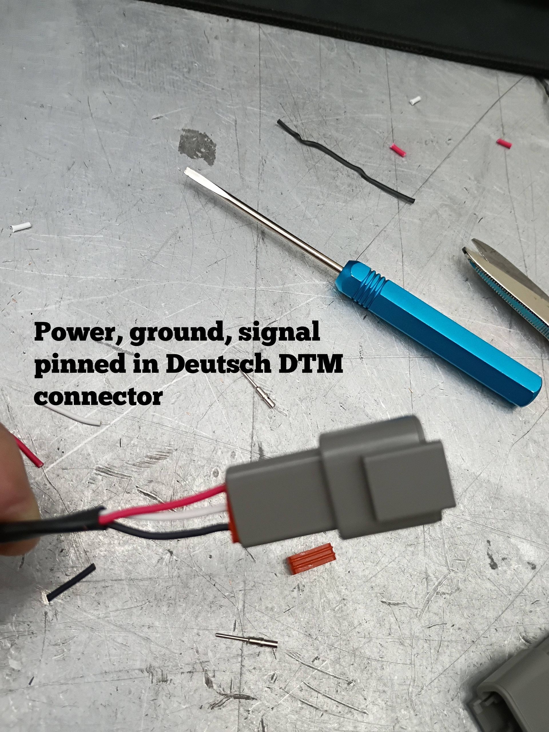 Gray Deutsch DTM connector with red and black wires on a workbench, labeled power, ground, and signal pinned in.